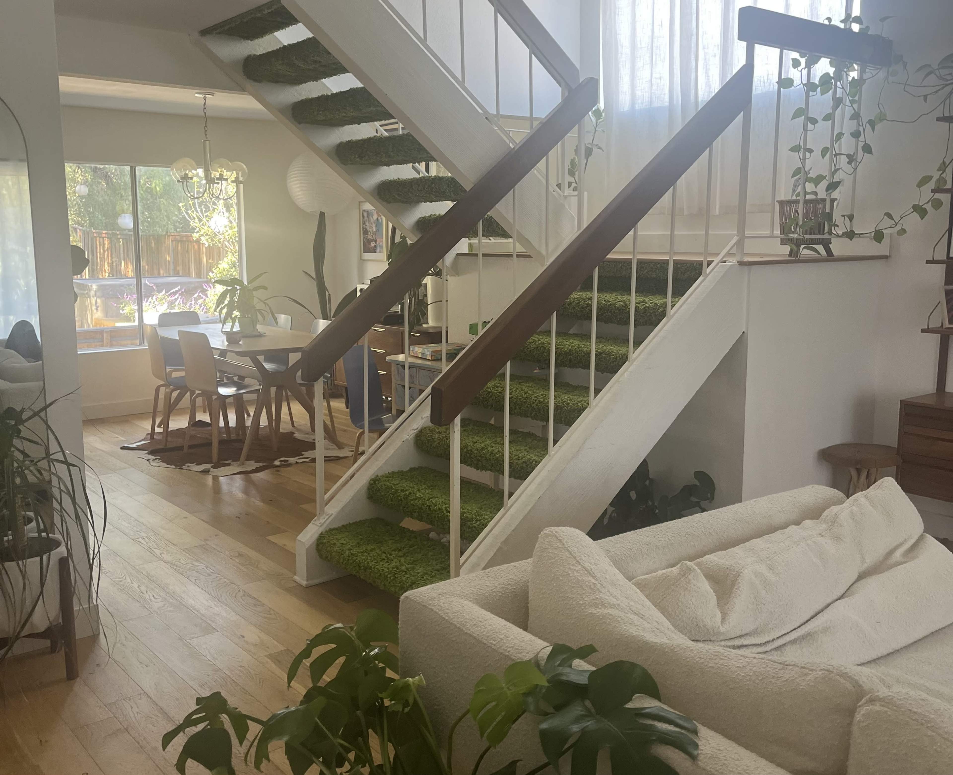 A bright, open living space featuring a staircase with green carpet, wooden flooring, and a cozy seating area surrounded by plants.