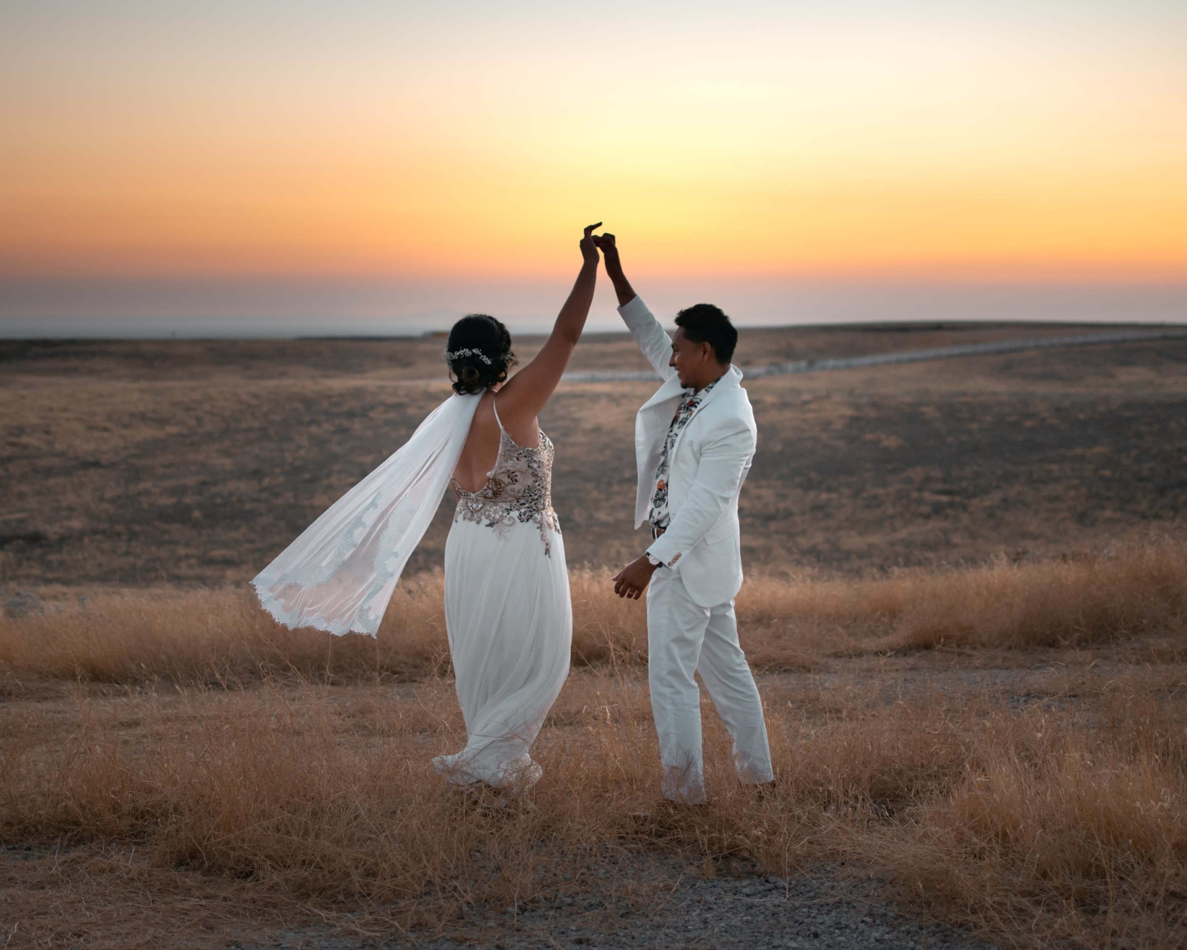 A couple in wedding attire dances together at sunset on a grassy hillside.