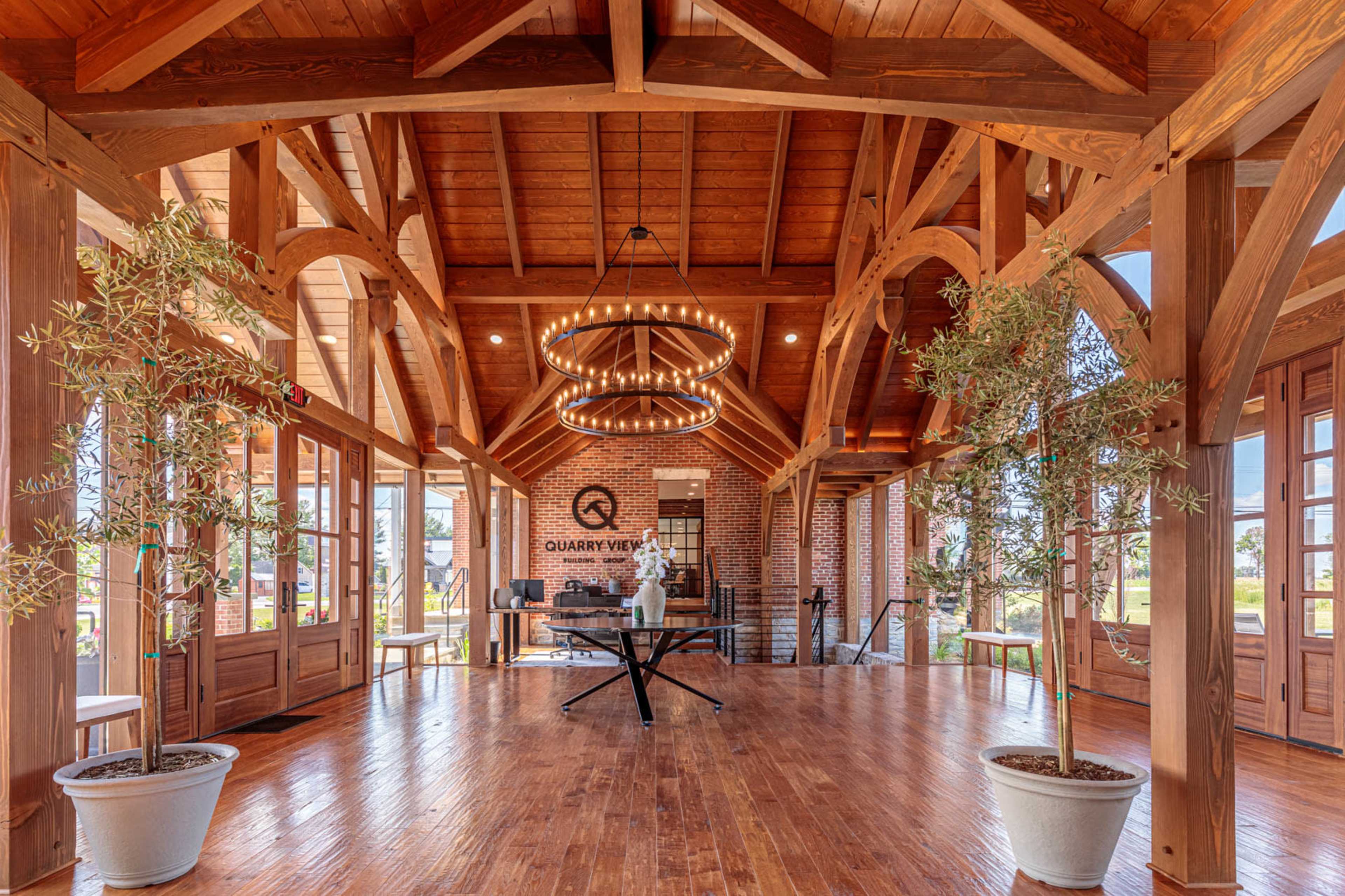The interior of a spacious wooden building features high ceilings with exposed beams, large windows, and potted plants arranged around a central table.
