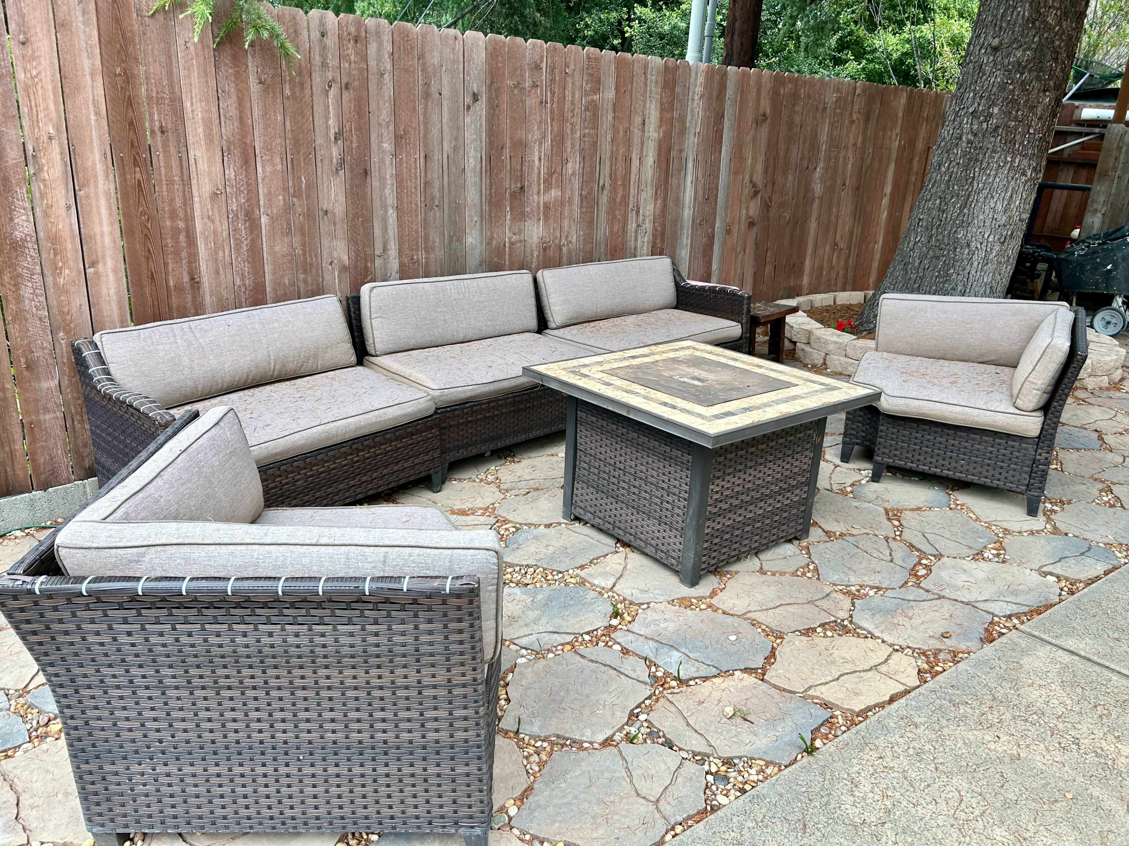 A sectional outdoor seating arrangement and a coffee table are positioned on a stone patio with a wooden fence and a tree in the background.