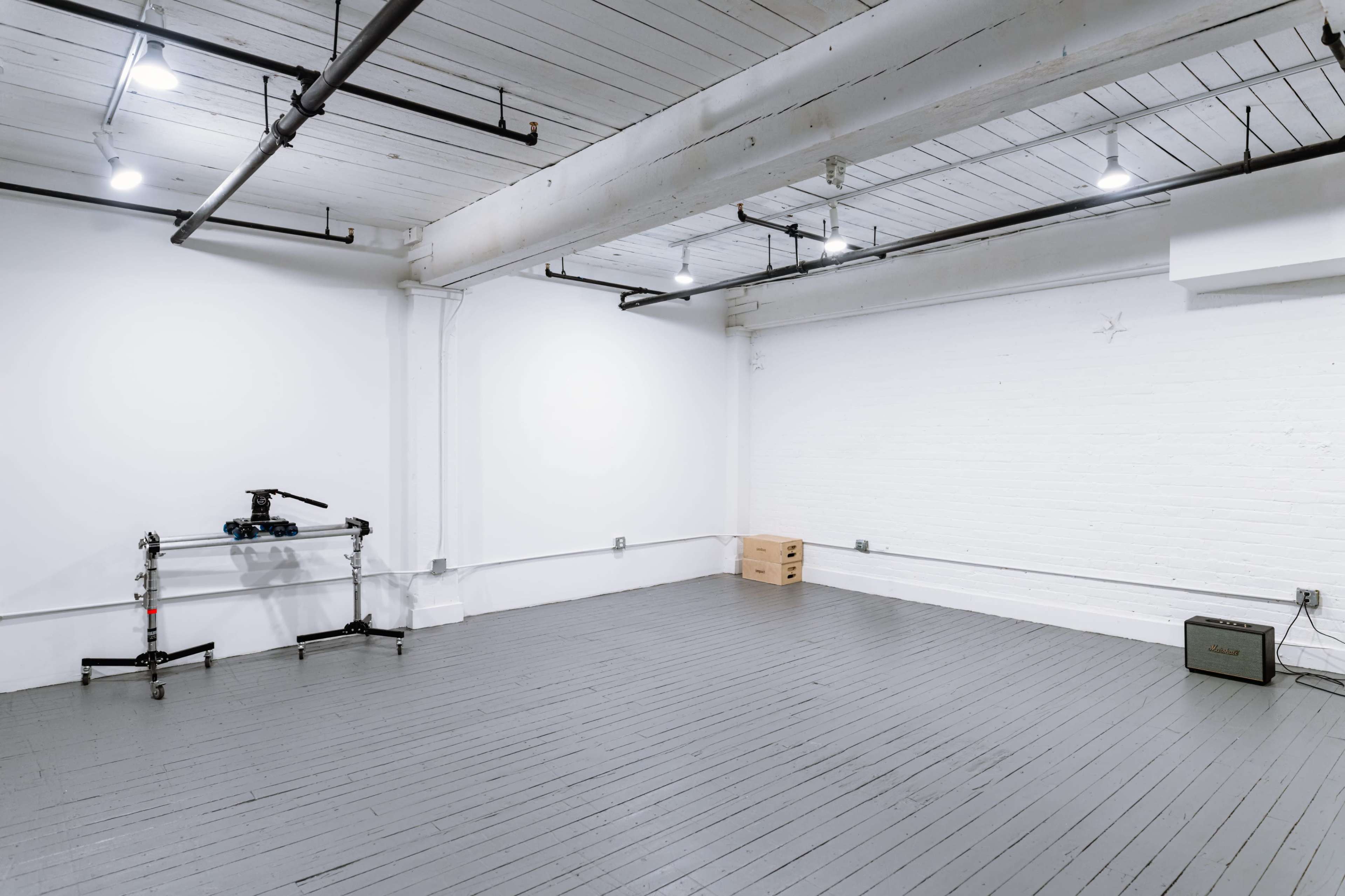 The image shows a spacious, empty room with light wooden floors, white walls, and a few lighting fixtures on the ceiling, along with a camera setup and boxes in one corner.