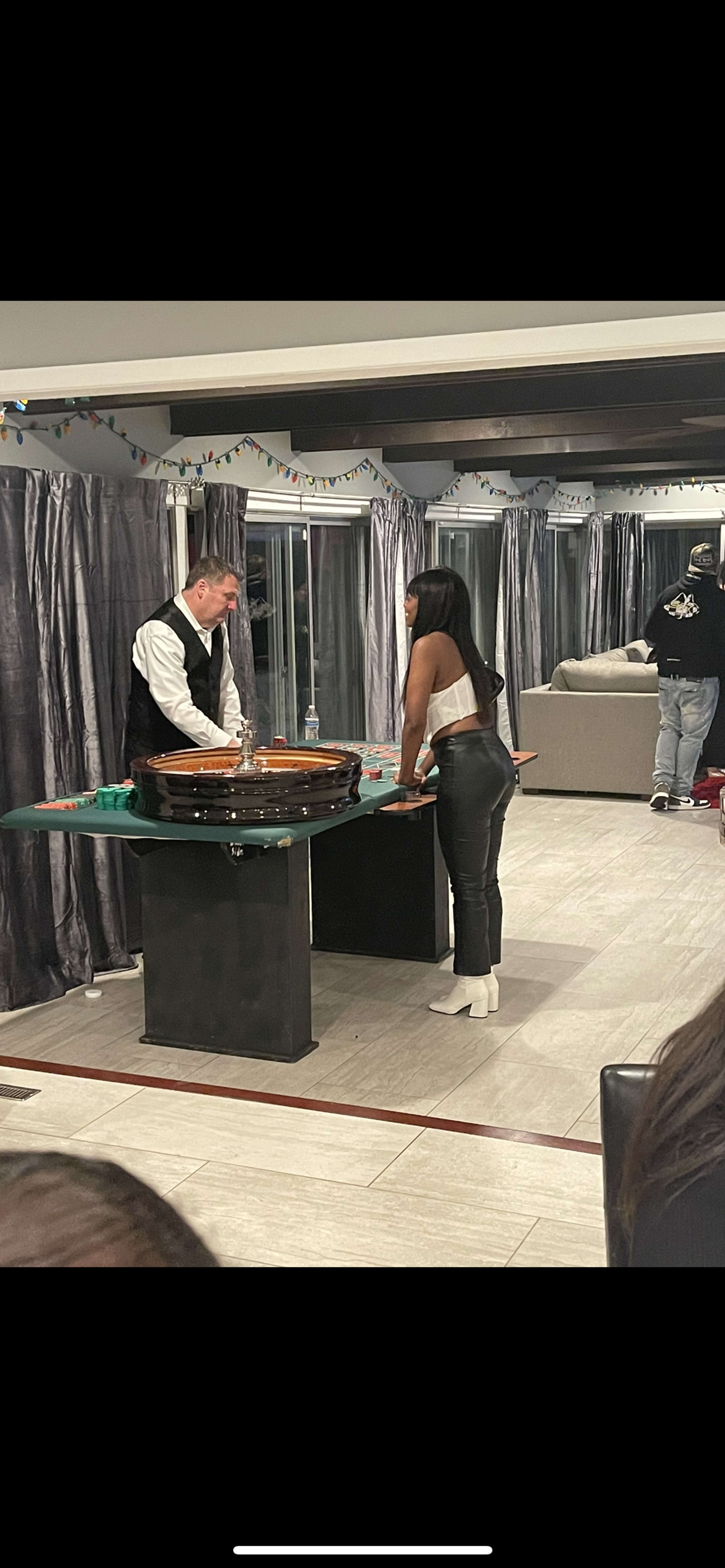 A dealer and a player engage in a game at a casino-style table in a spacious room with curtains and festive decorations.