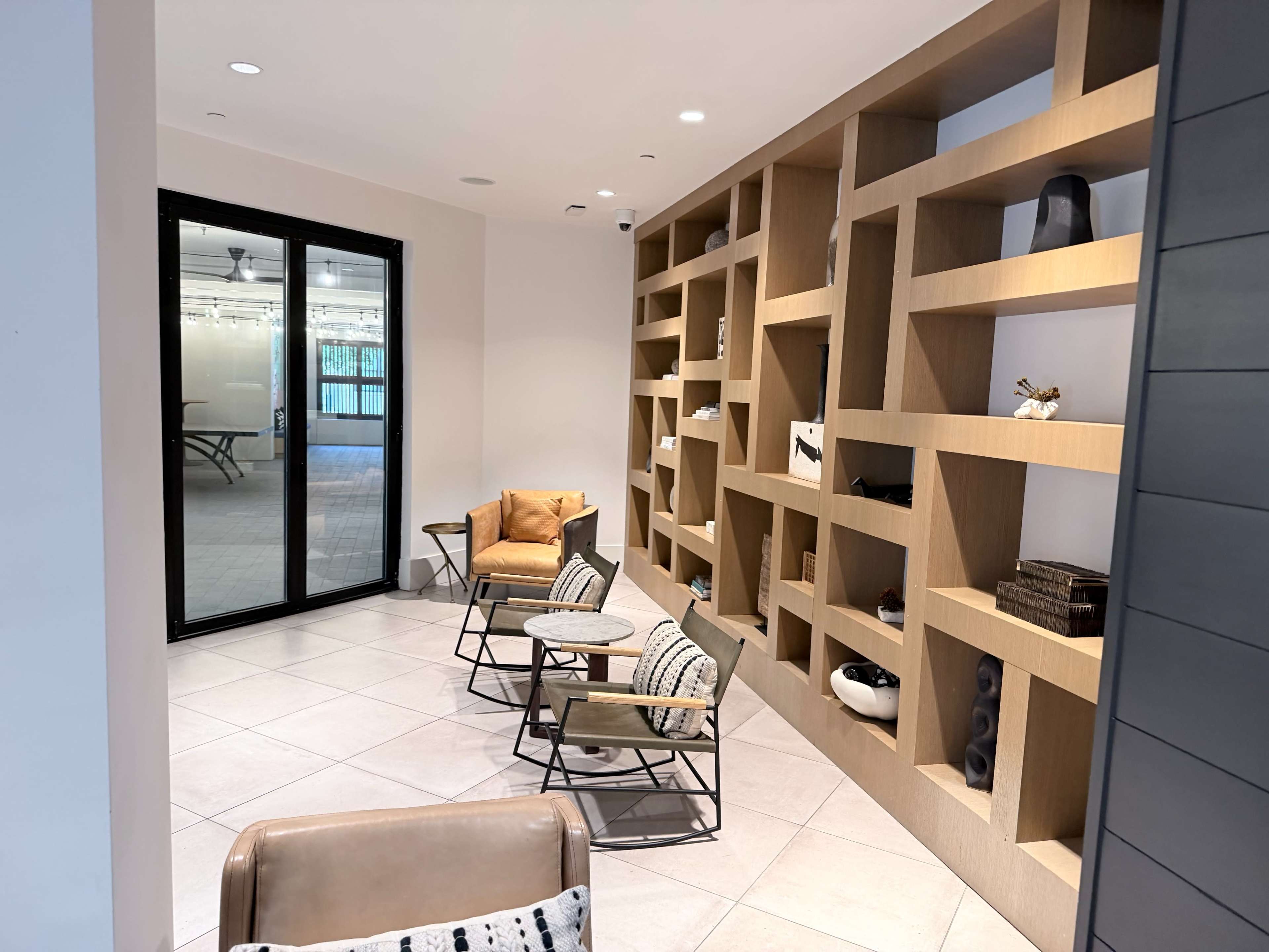The image shows a cozy seating area with two chairs and a small table in front of a modern shelf unit, adjacent to a glass door leading to another room.