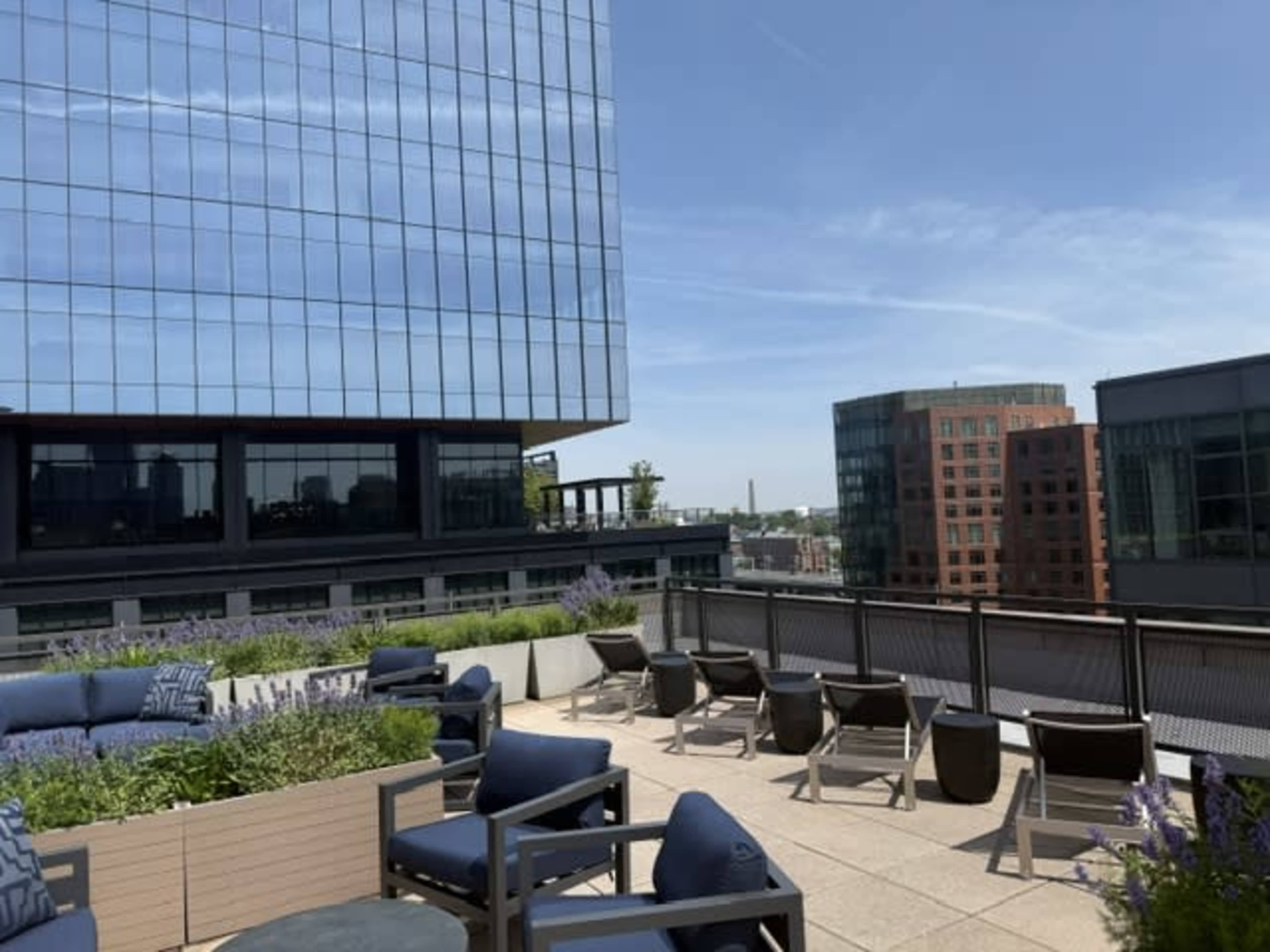 A rooftop terrace features seating arrangements, modern furniture, and greenery, with a view of surrounding buildings under a clear blue sky.