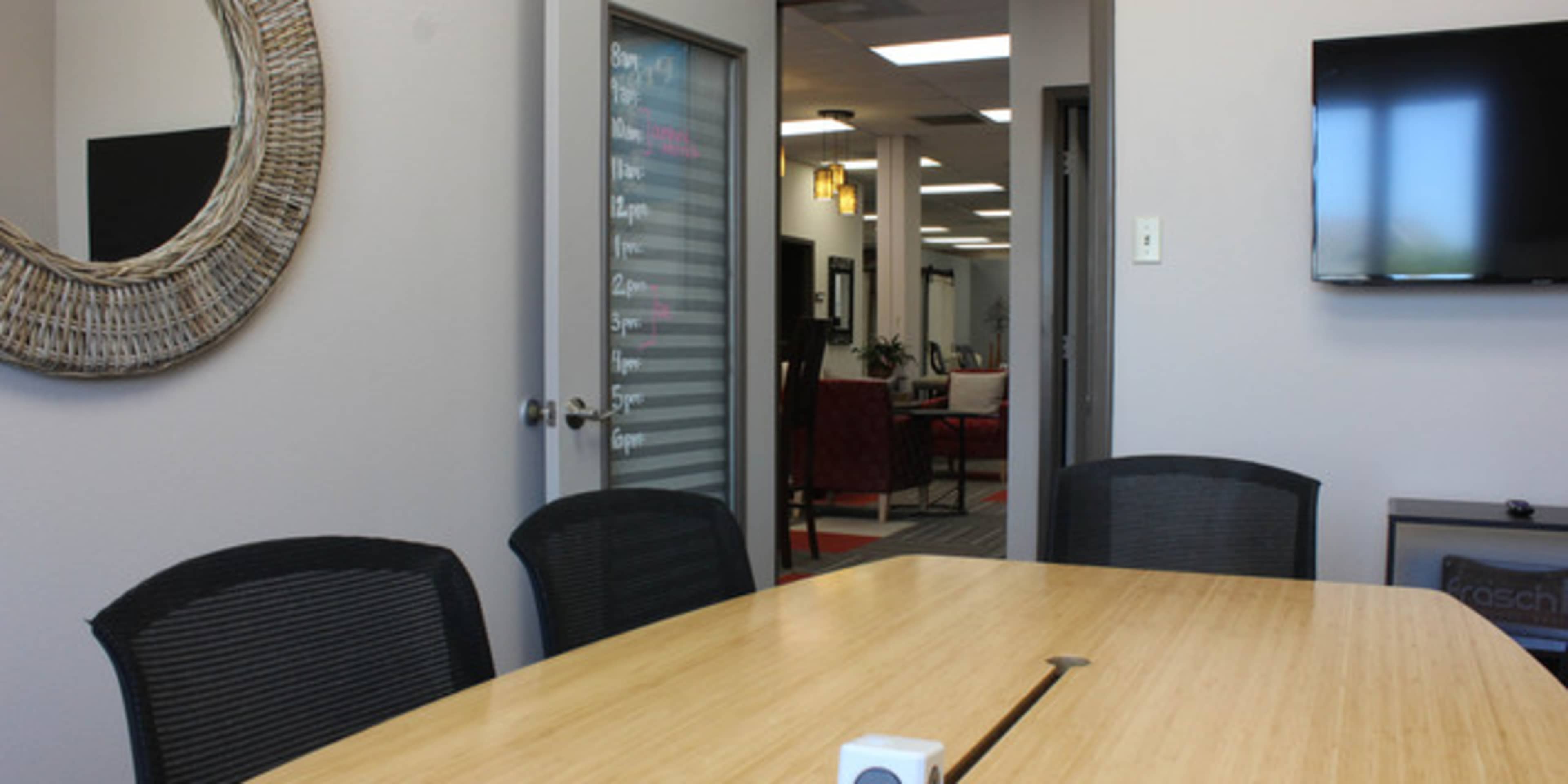 A conference room features a wooden table surrounded by black chairs, with a large mirror and a television mounted on the wall.