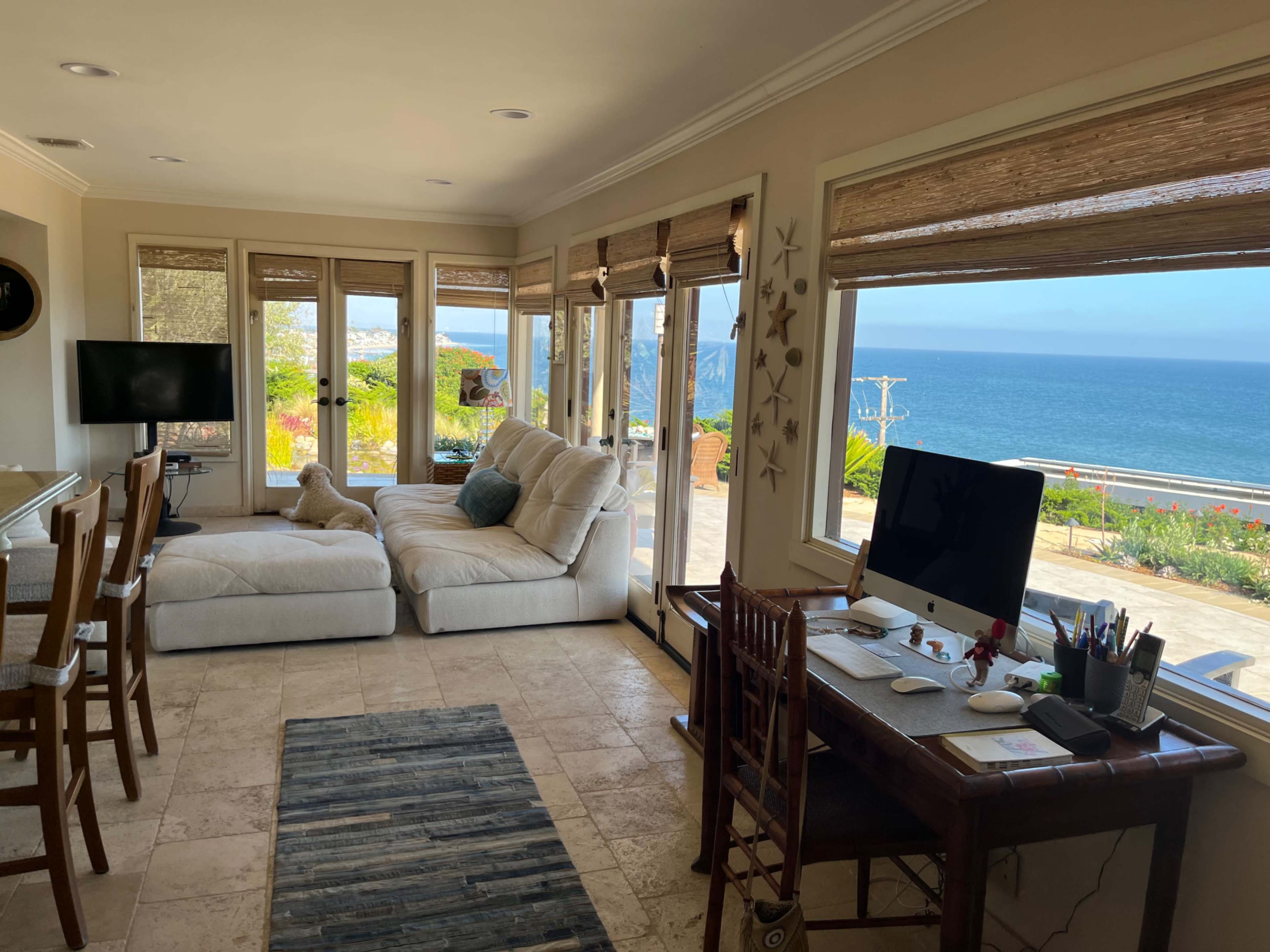 A bright living room features large windows overlooking the ocean, with a comfortable sofa, a desk with office supplies, and wooden dining chairs.