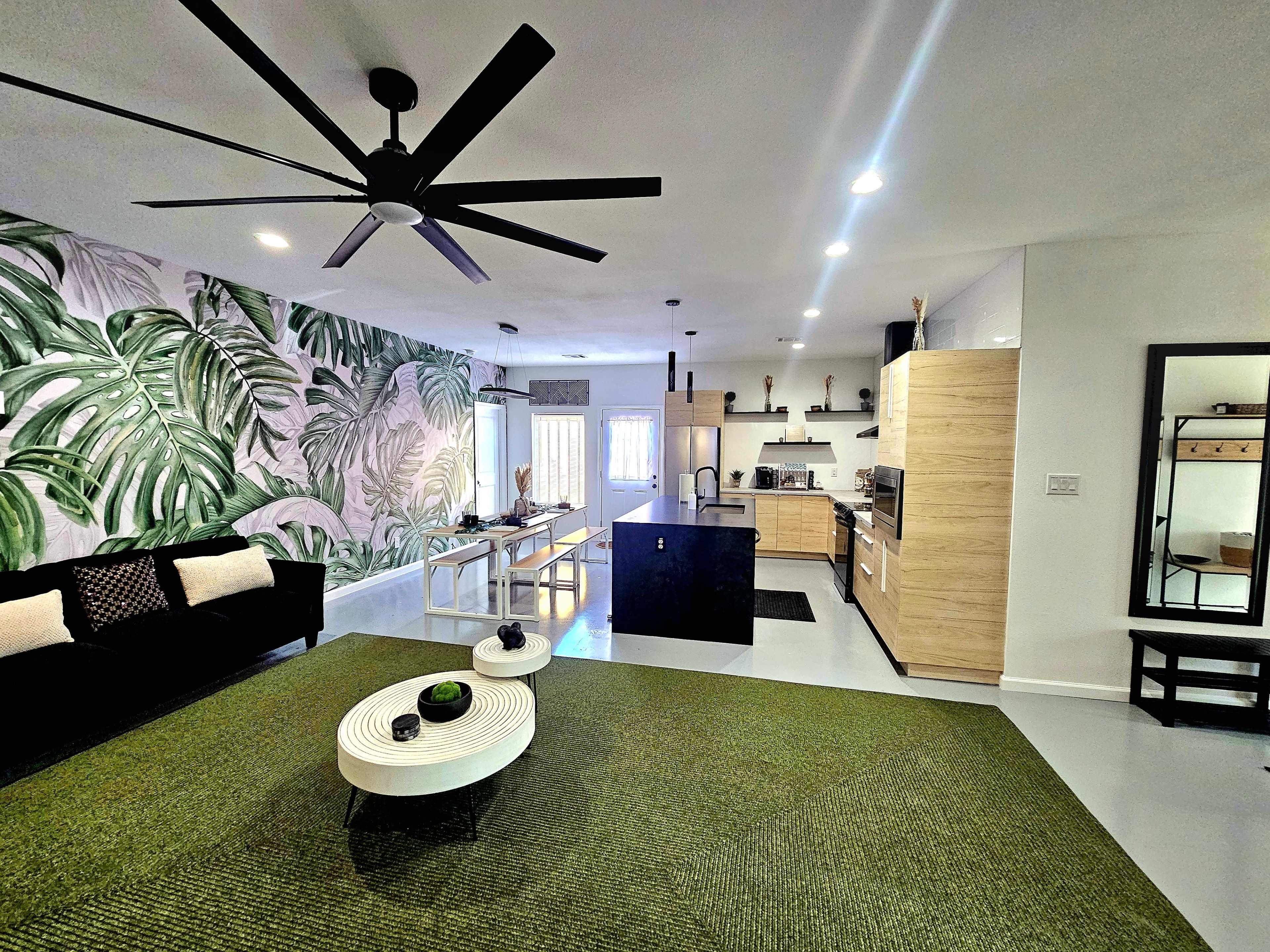A spacious living area featuring a dark sofa, a round coffee table, a large green area rug, and a kitchen with light-colored cabinetry and a tropical leaf mural on the wall.