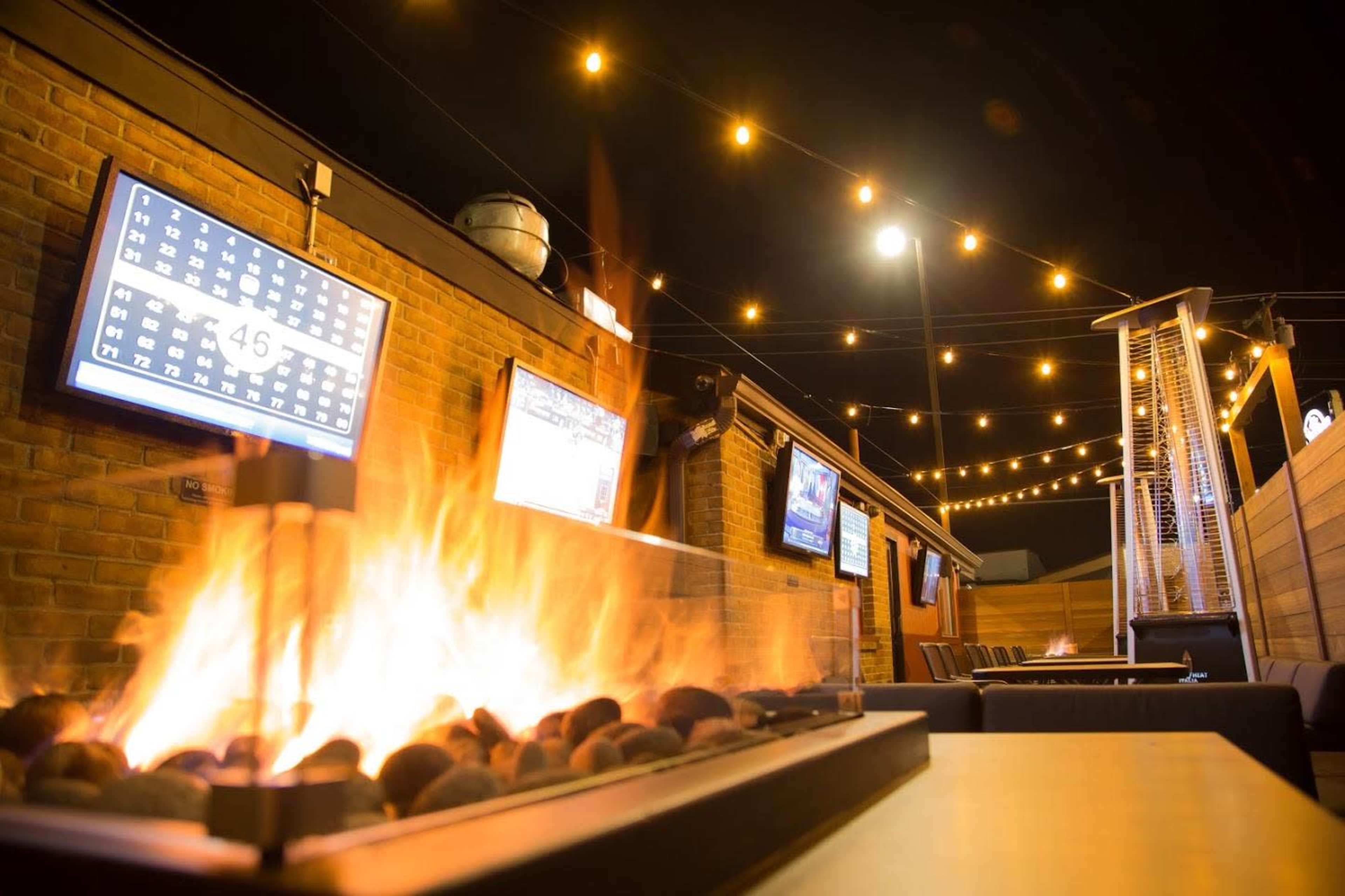 An outdoor seating area with a central fire feature, several TVs mounted on the walls, and string lights overhead.