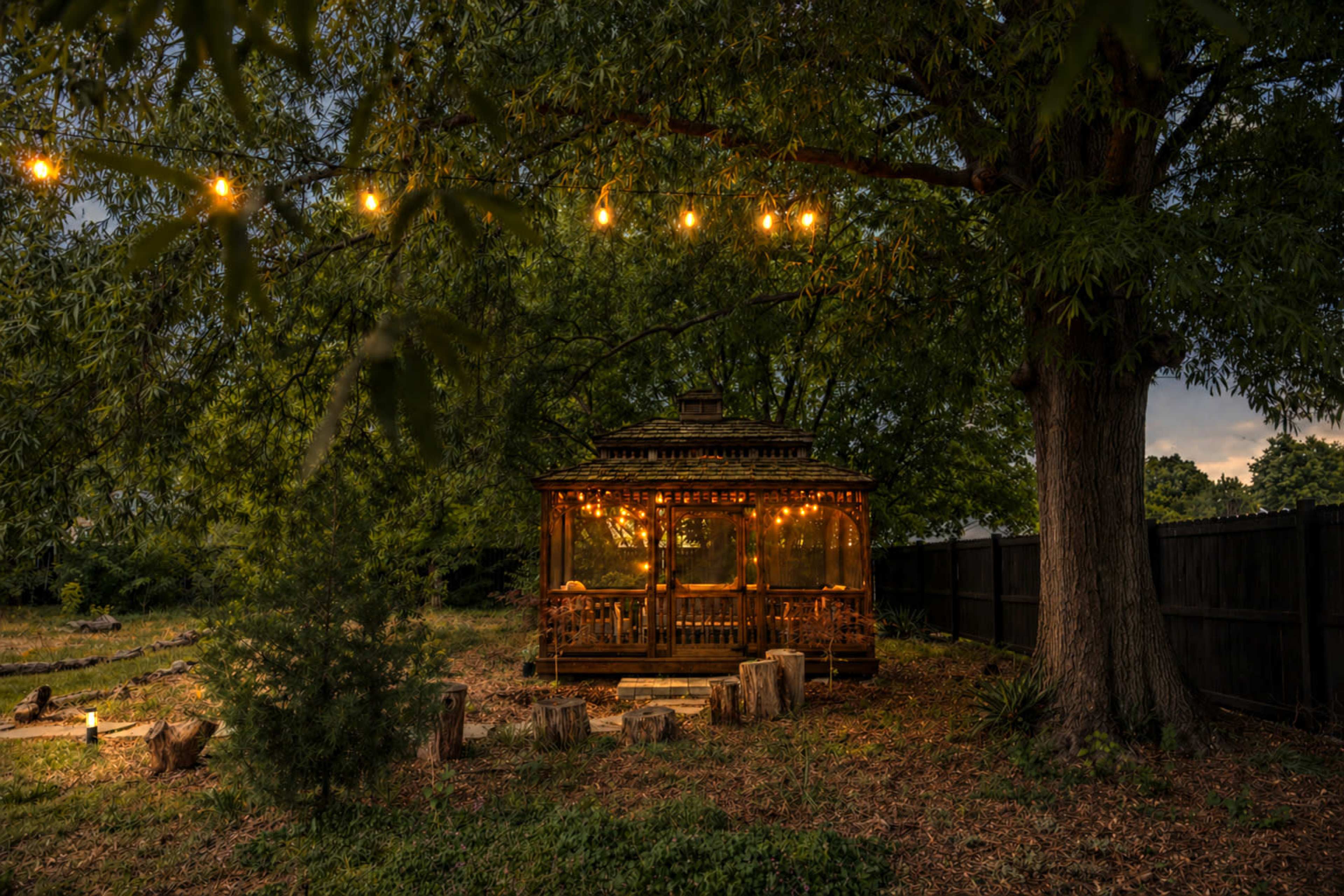 A wooden gazebo adorned with string lights sits in a grassy yard under trees.