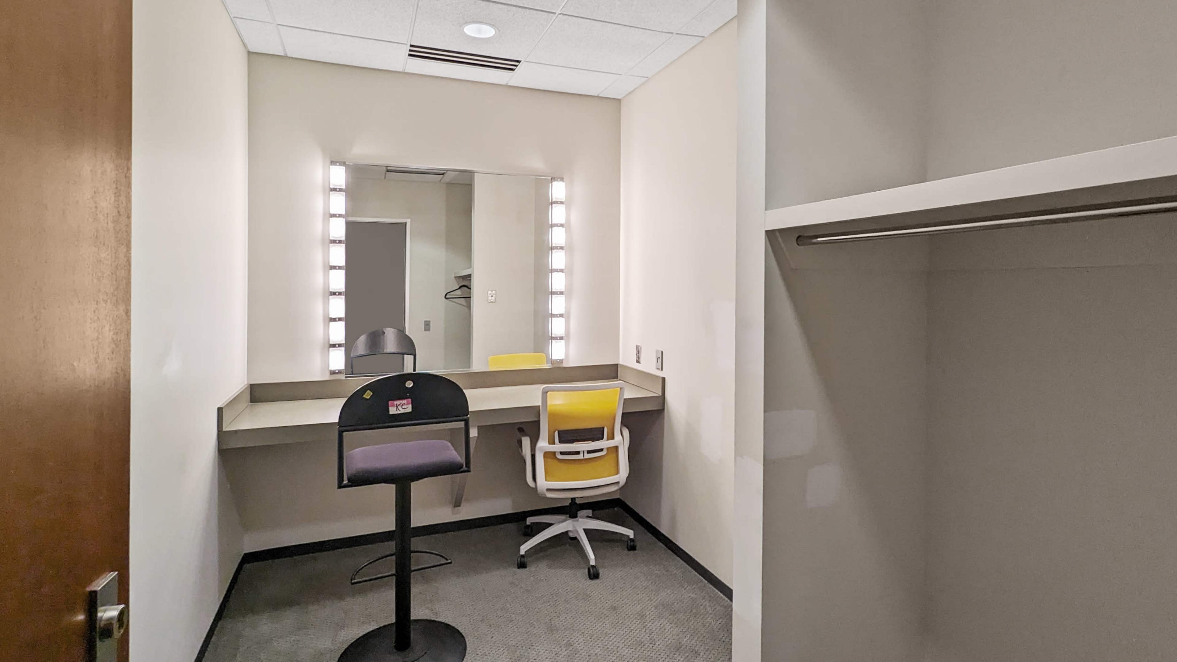 The image shows a simple dressing room with a mirror, lighted on the sides, a chair, and an empty closet space.