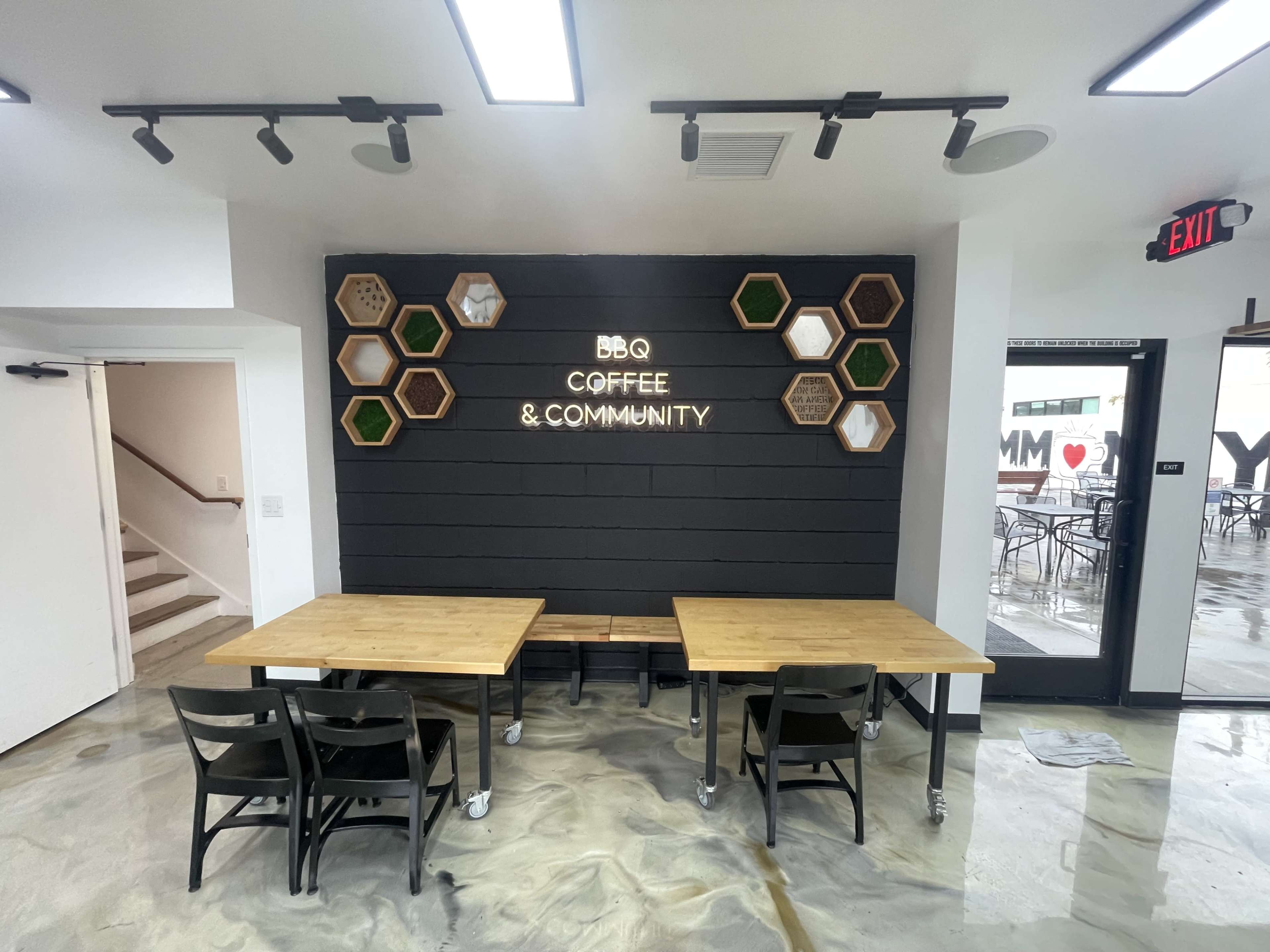 The image shows a modern coffee shop interior featuring a wall with hexagonal wood and green wall decor, and two wooden tables with black chairs.