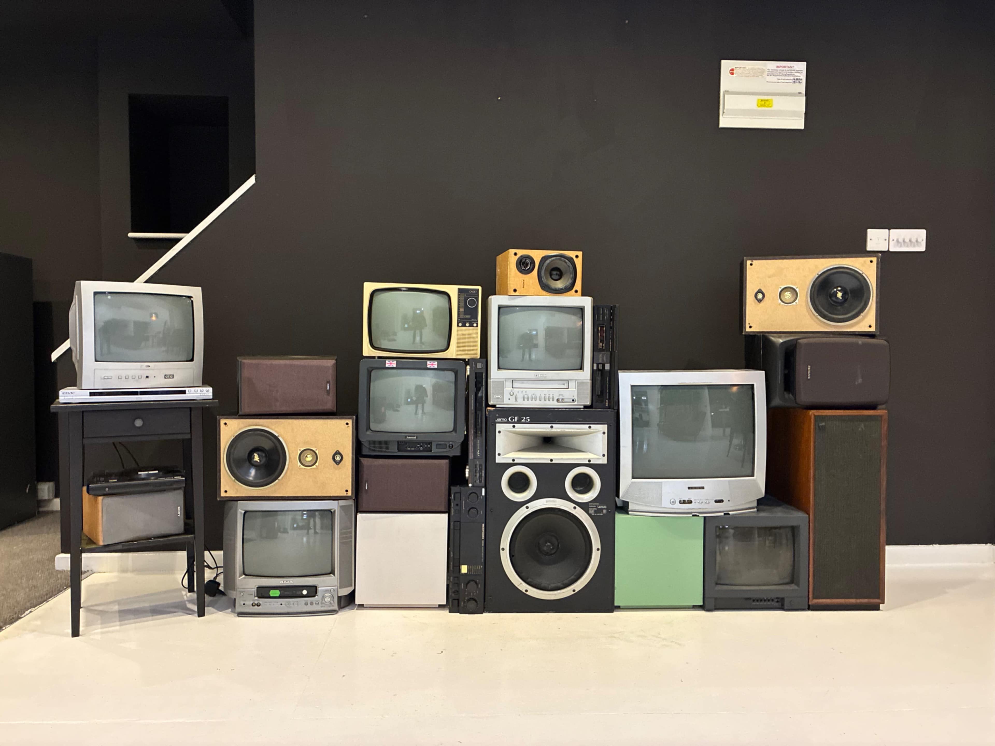 The image shows a collection of vintage televisions and audio equipment arranged in a stacked formation against a black wall.
