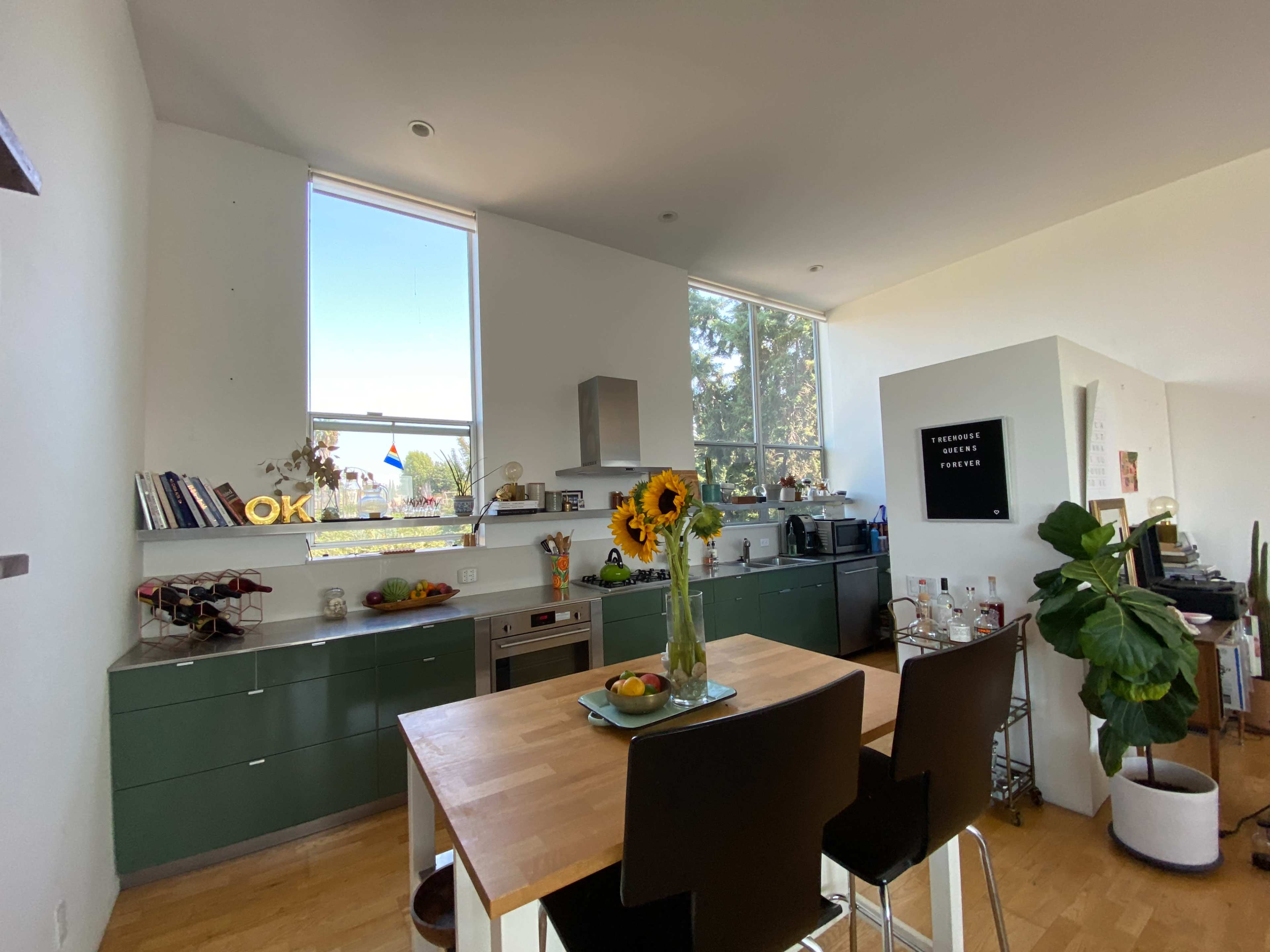 A modern kitchen features a wooden table with two black chairs, green cabinetry, and large windows that provide natural light and a view.