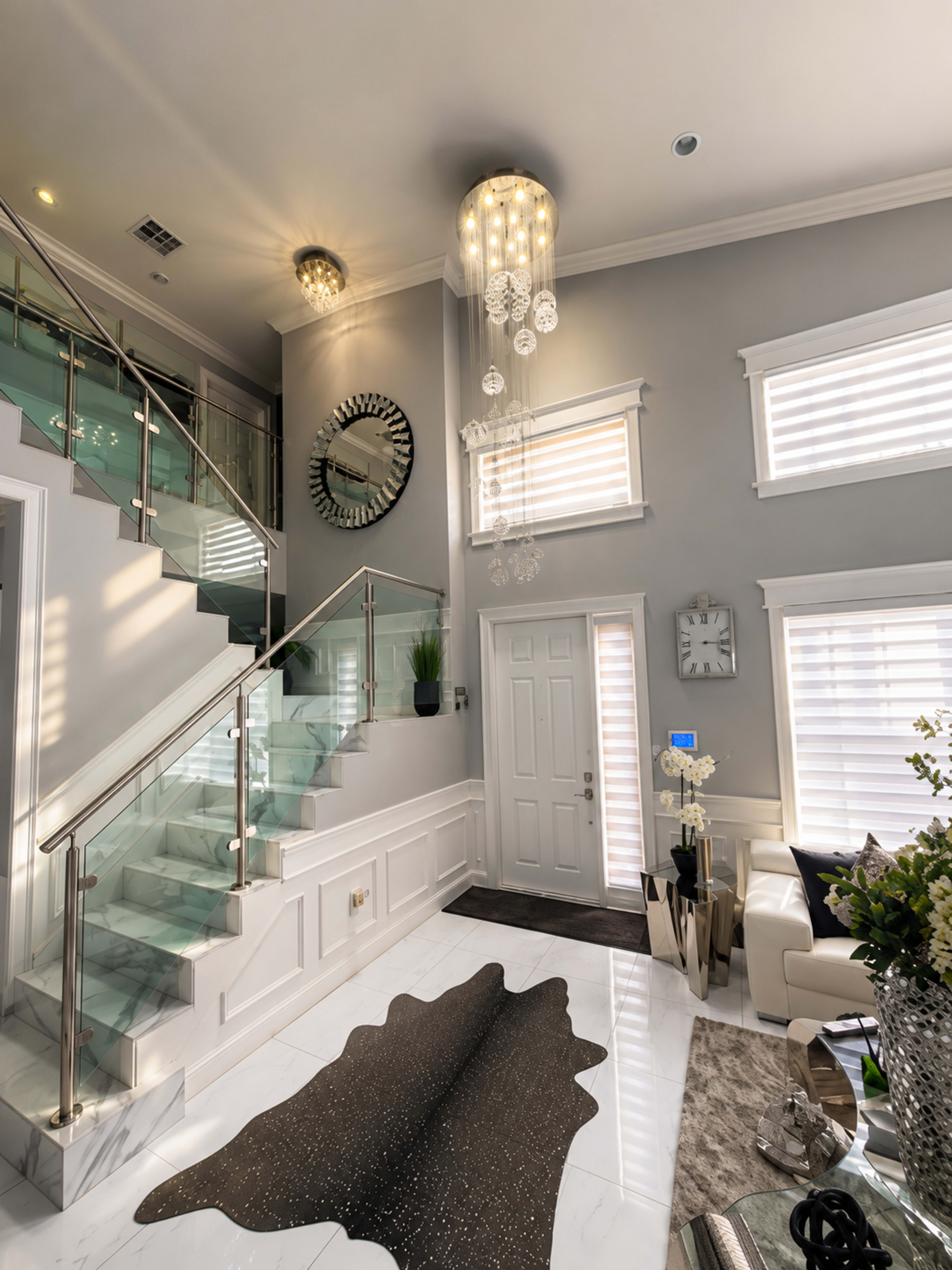 The image shows a modern foyer with a glass staircase, a large circular mirror, a crystal chandelier, and elegant furnishings, including a black-and-white rug and a decorative plant.