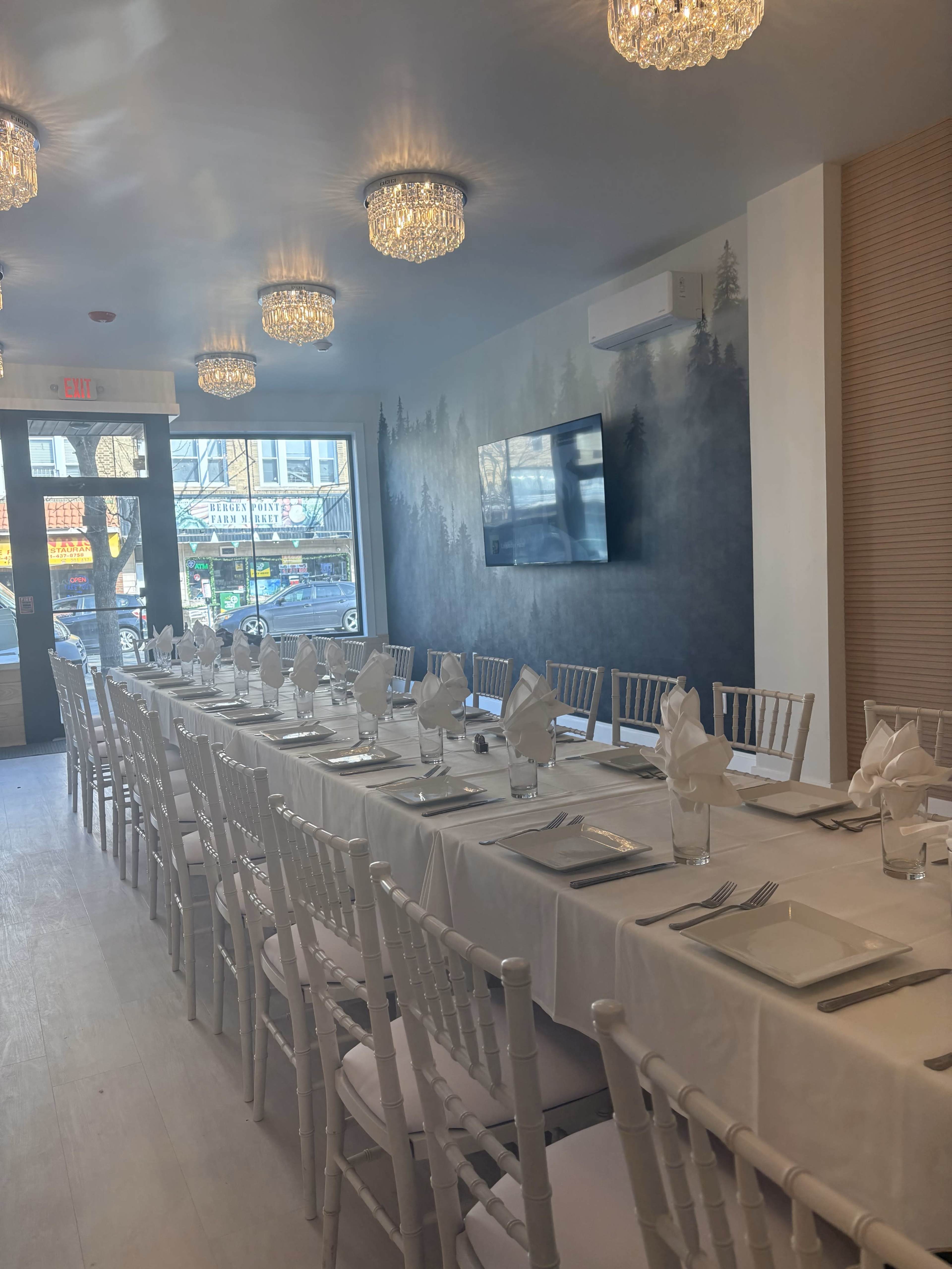 The image shows a long banquet table set with white linens, plates, and napkins, arranged inside a well-lit restaurant featuring wall decor and a television.