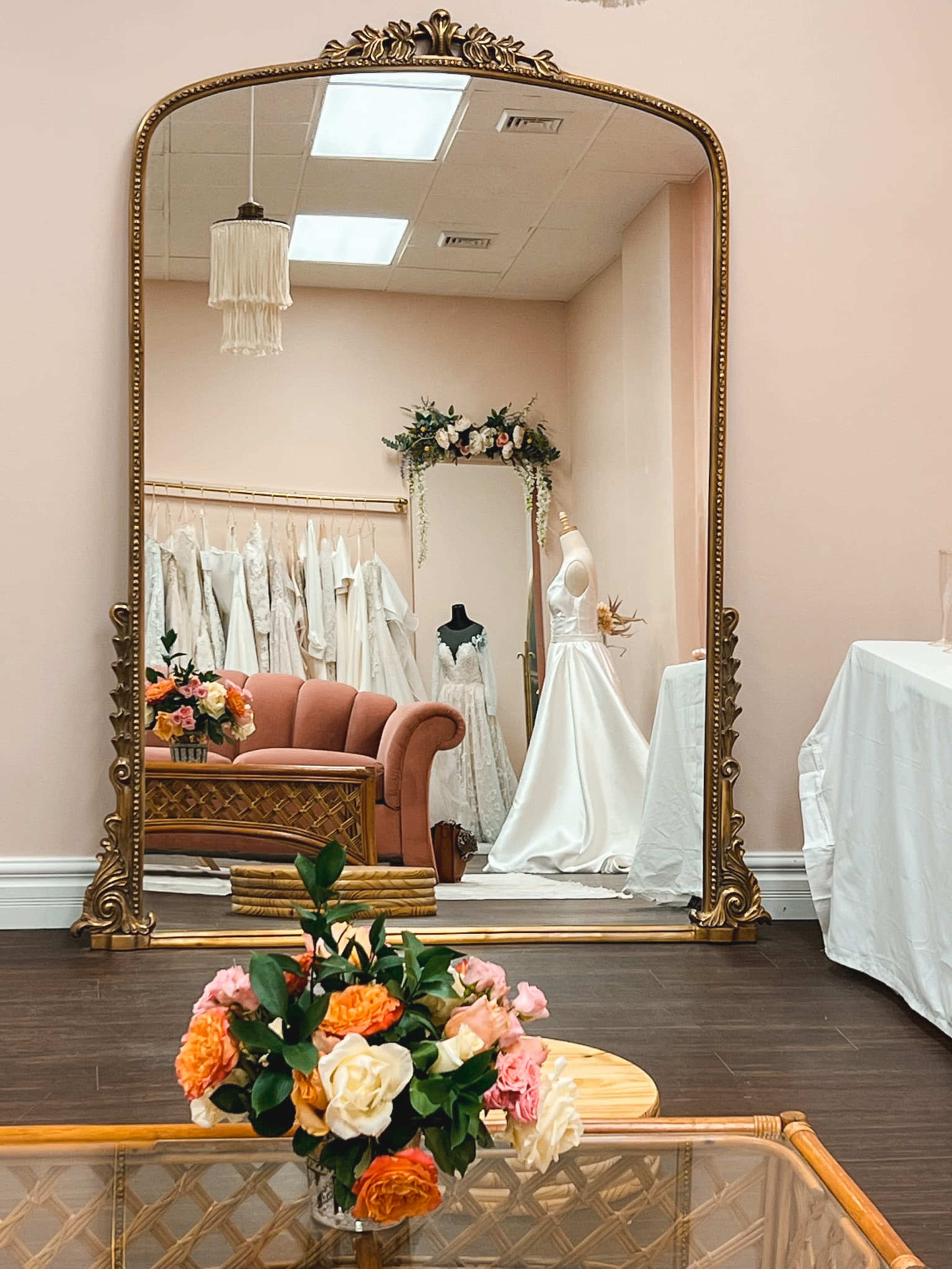 A large ornate mirror reflects a seating area with a pink couch and displays of wedding dresses in a boutique setting.