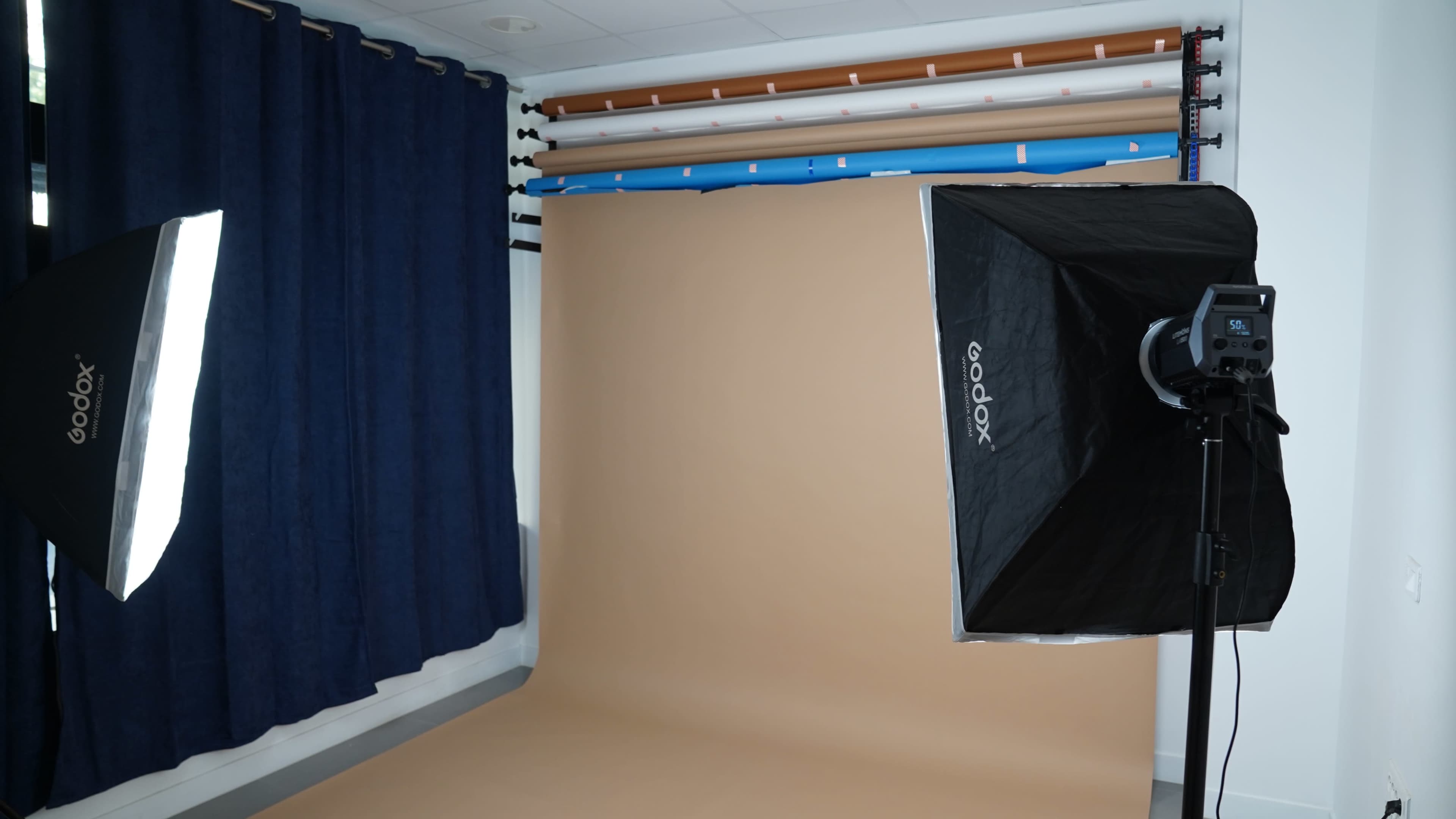 The image shows a photography studio featuring a large softbox light, a seamless background roll system with a tan backdrop, and dark curtains along one wall.