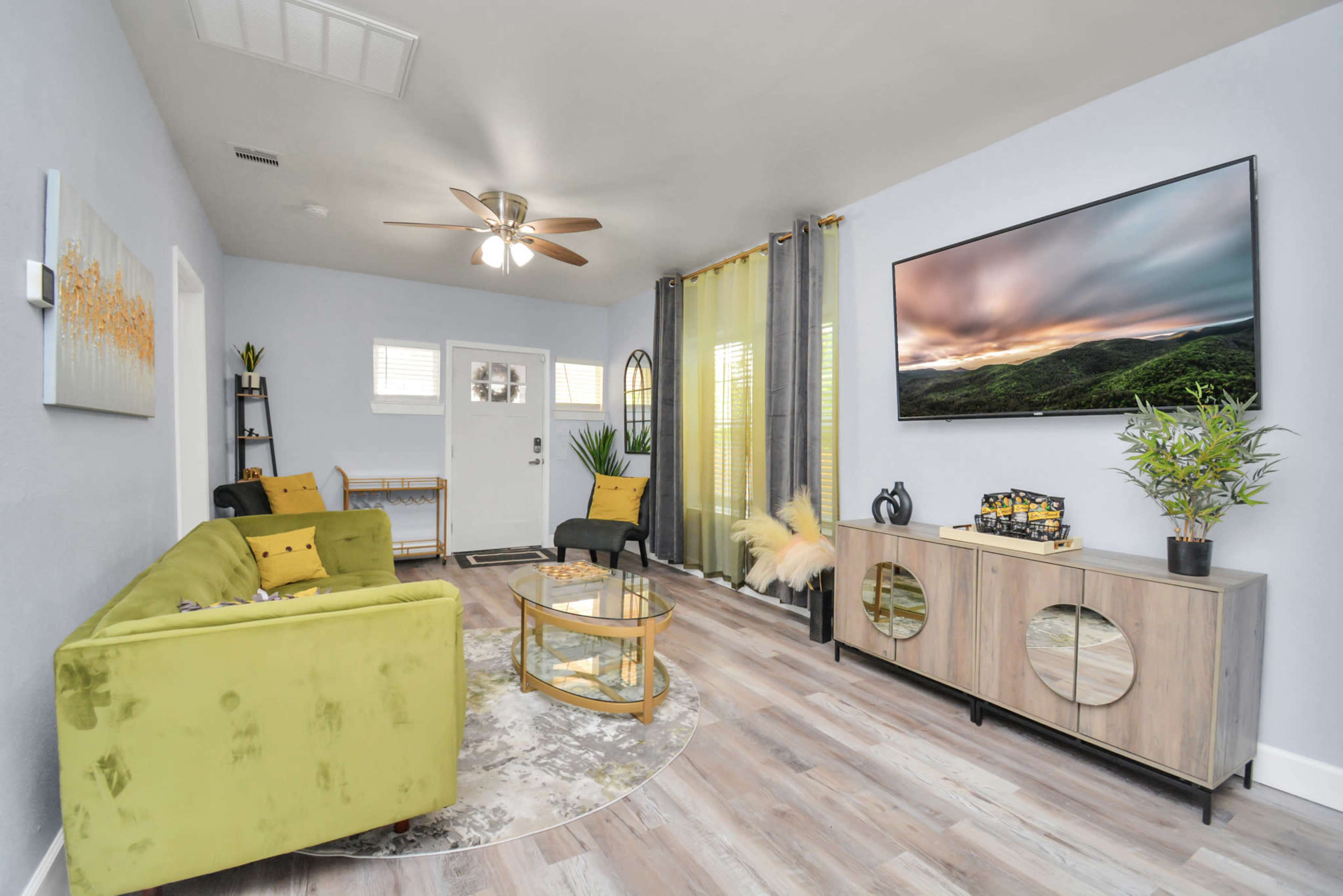 A modern living room with a green sofa, a round coffee table, a television mounted on the wall, and large windows with curtains.