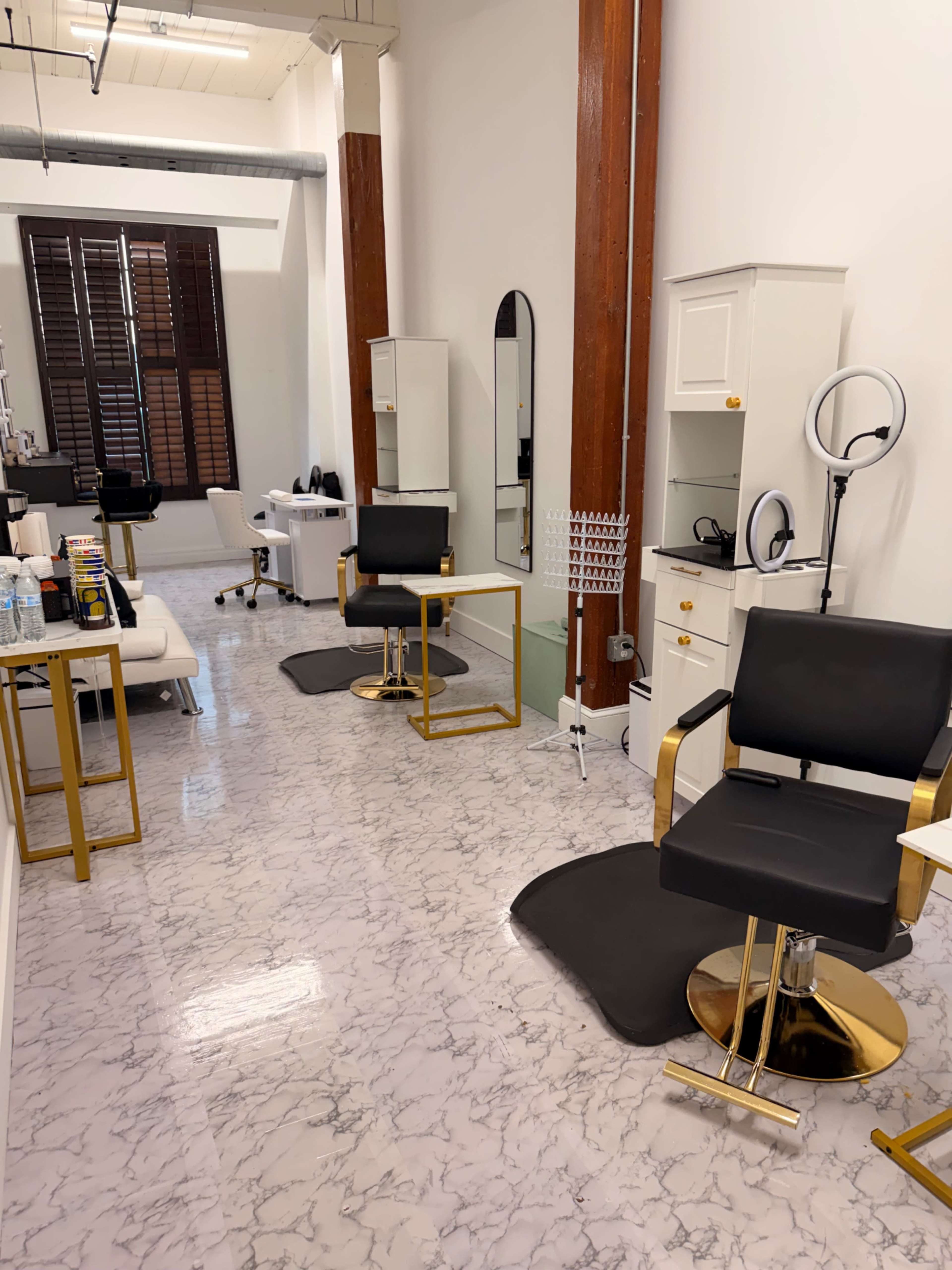 The image shows a modern salon interior with black salon chairs, a large mirror, and various styling tools on display in a spacious, well-lit area.