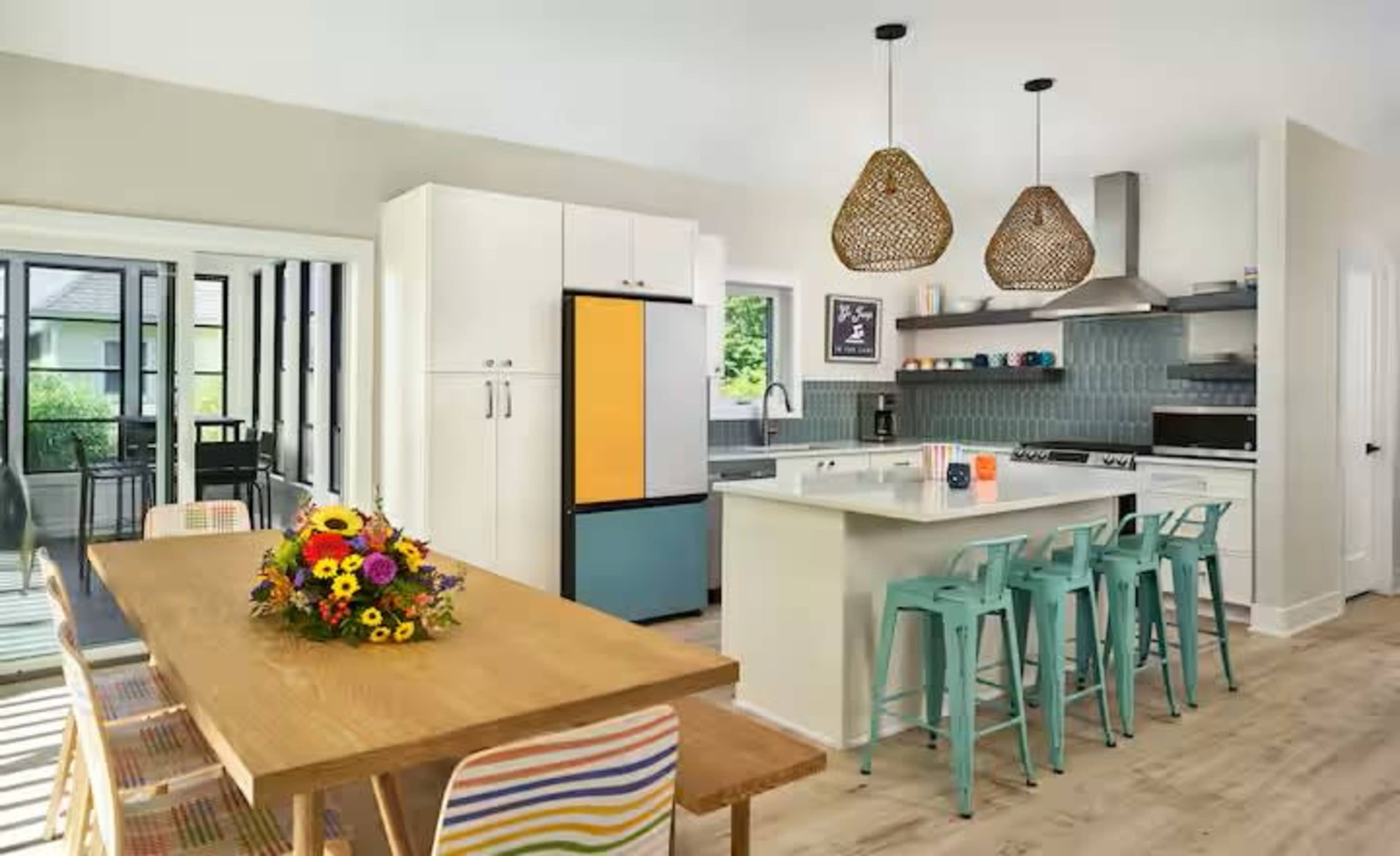The image shows a modern kitchen with a dining area, featuring a wooden table, colorful chairs, and pendant lighting above a kitchen island.