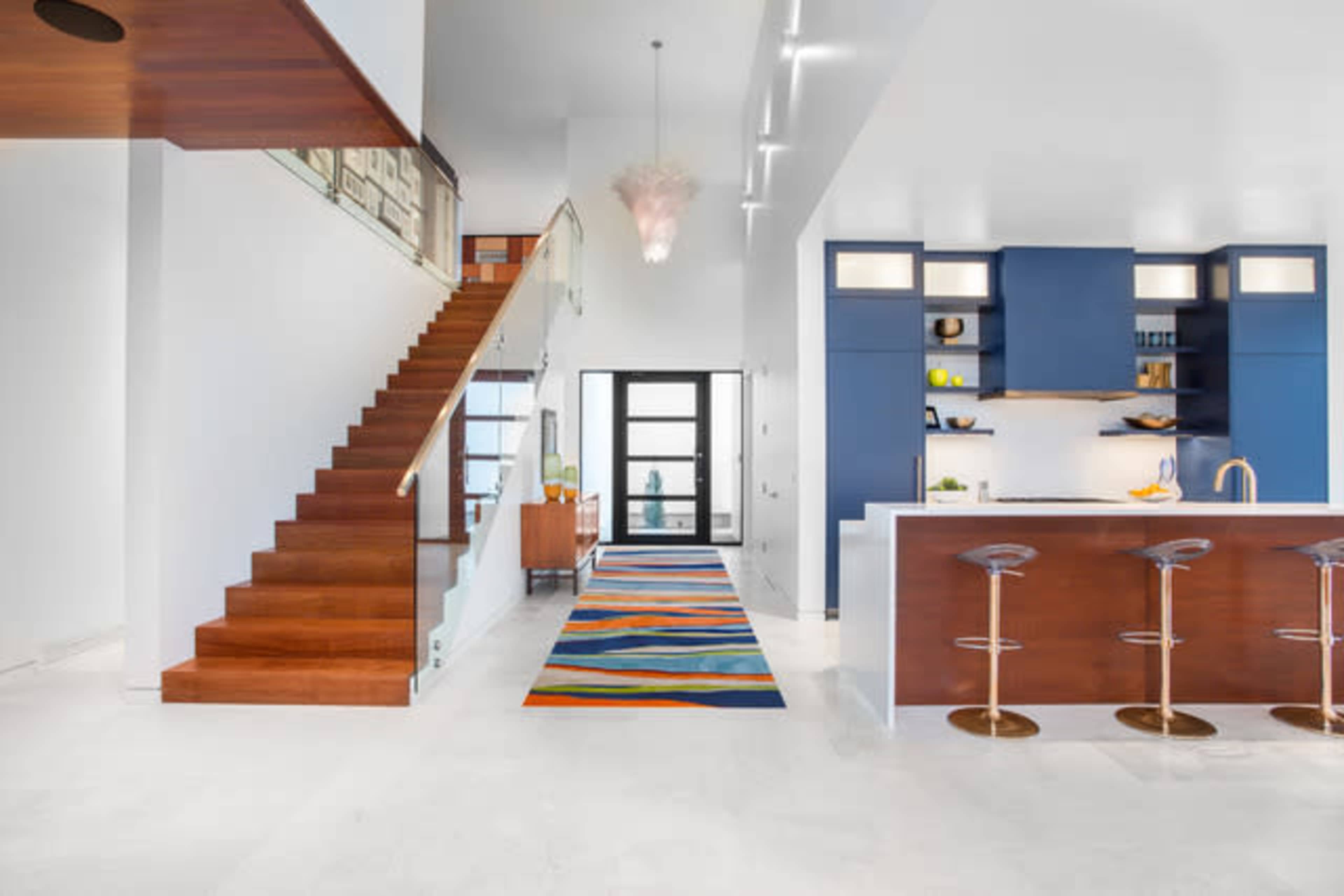 The image shows a modern interior with a staircase leading to an upper level, a kitchen island with bar stools, and colorful rugs on a light-colored floor.
