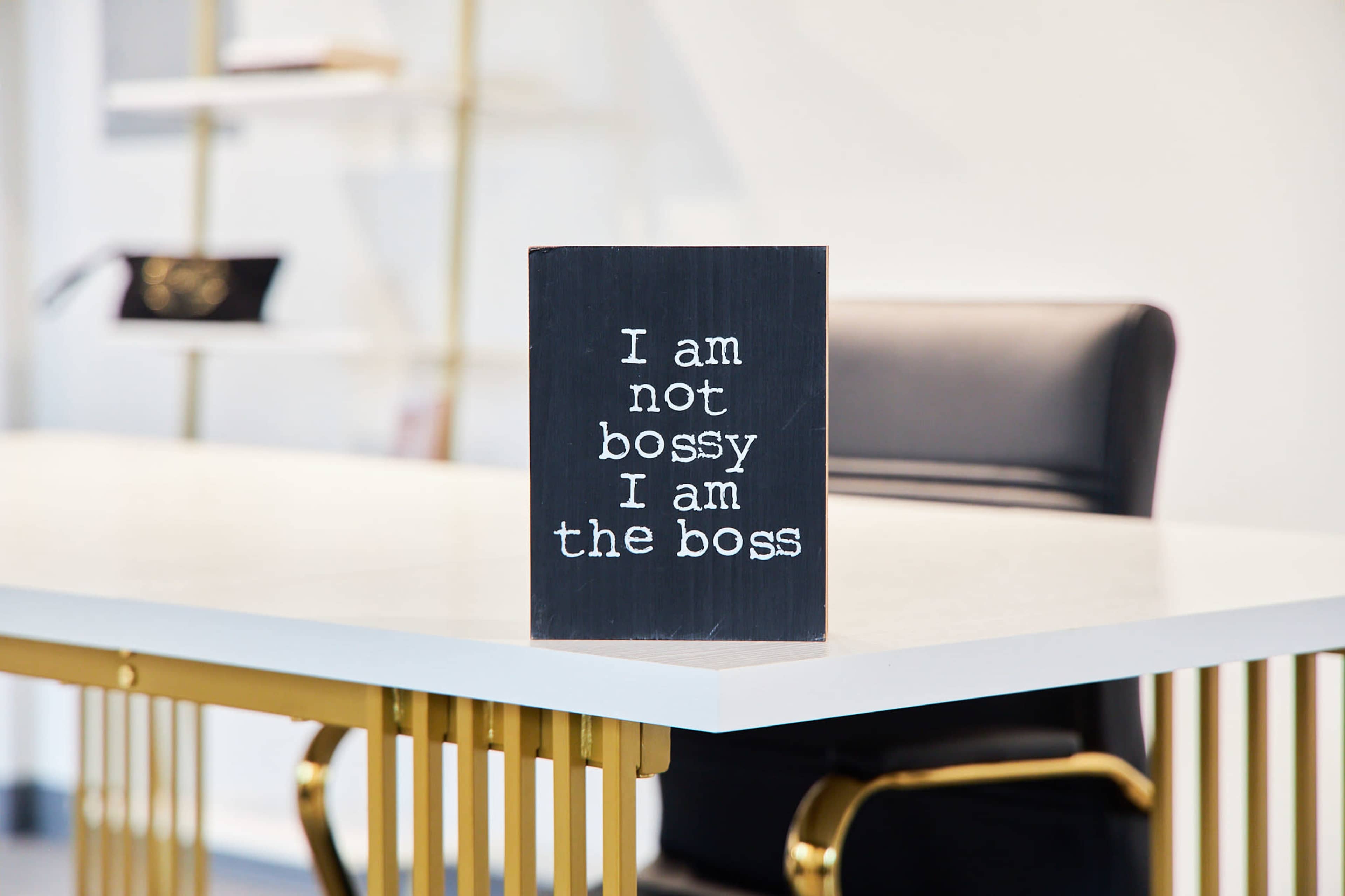A sign reading "I am not bossy I am the boss" is placed on a white desk in an office setting.