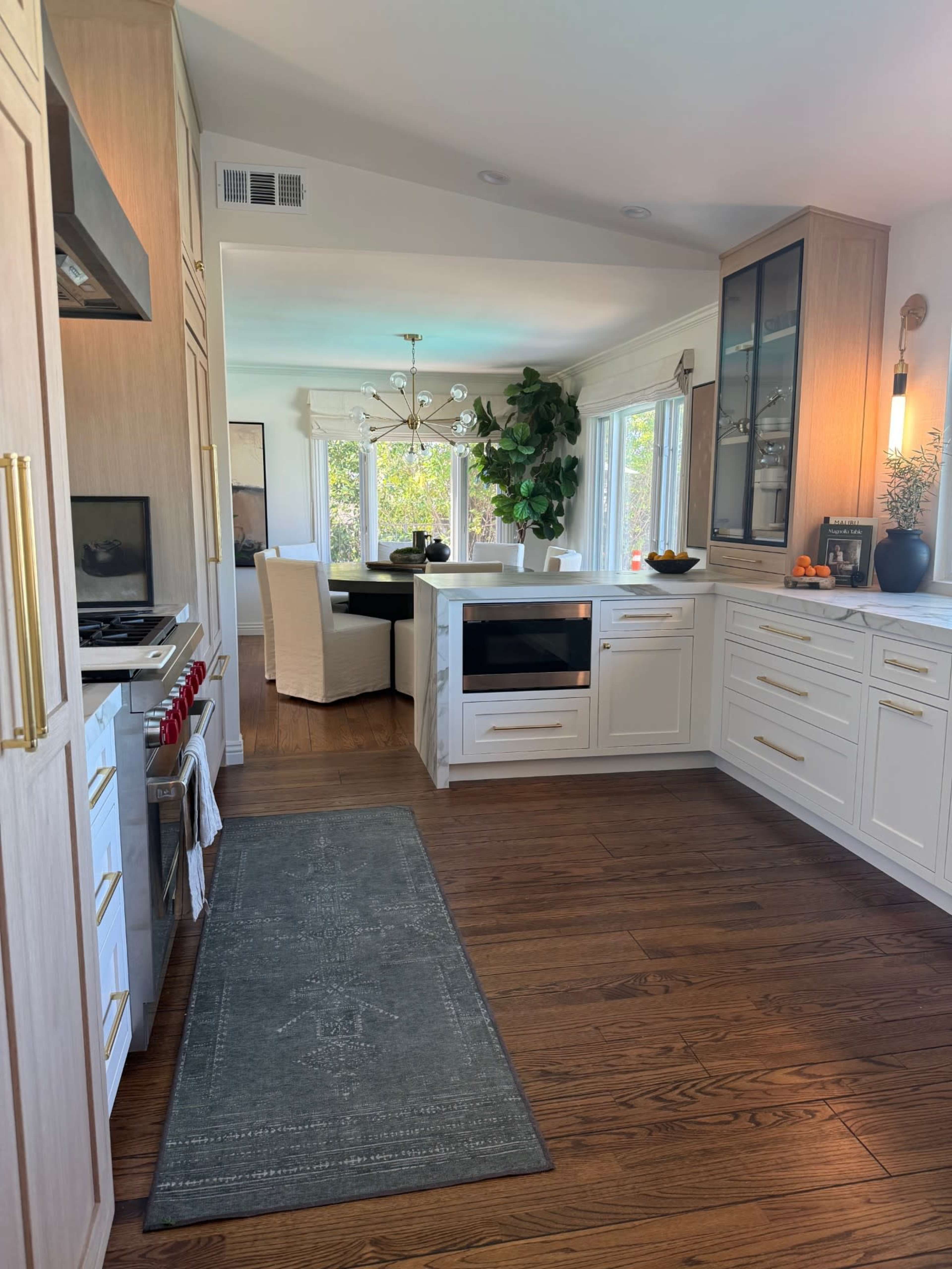 The image shows a modern kitchen with light-colored cabinets, a central island with a stovetop, and a dining area featuring white chairs and a large window.