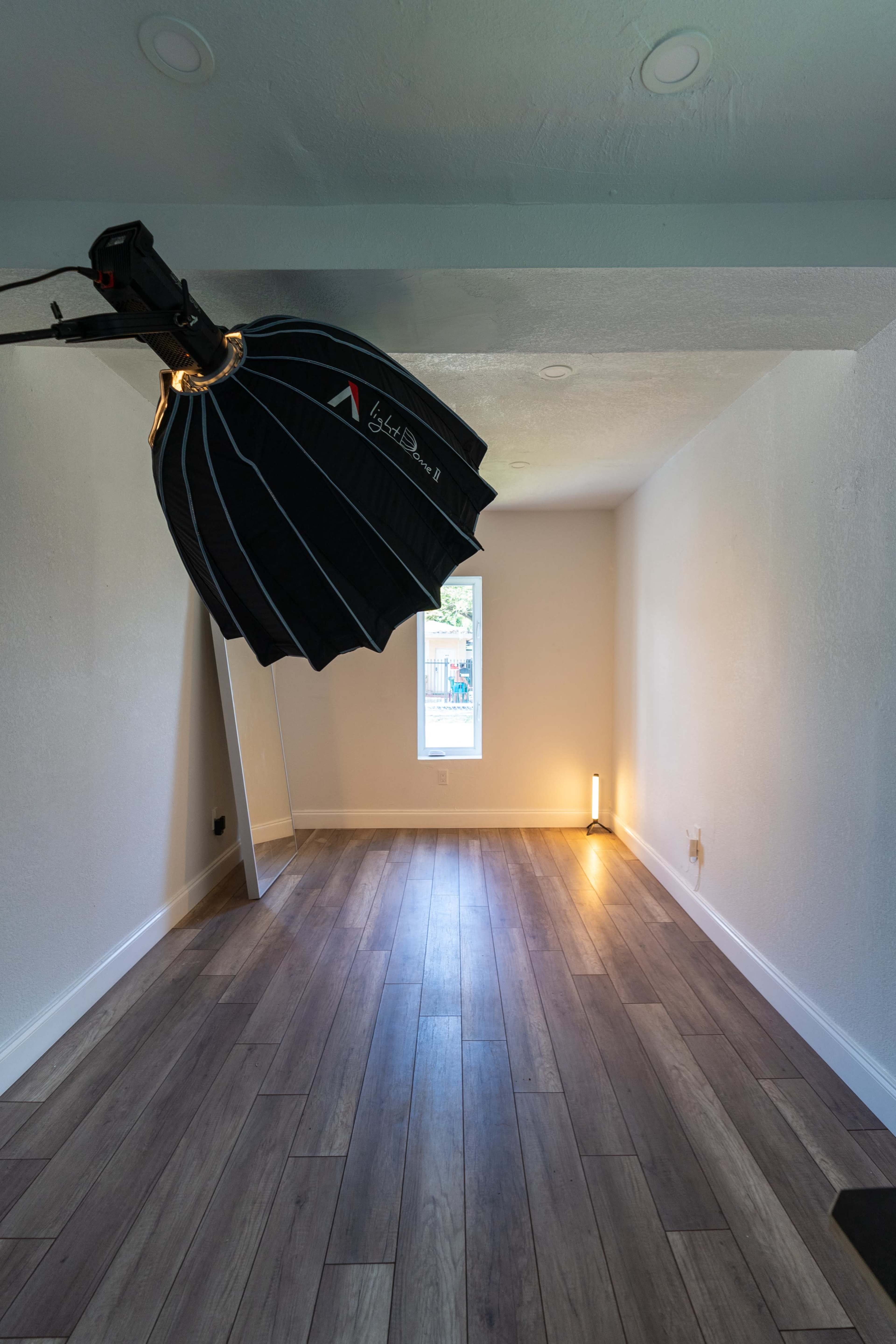 An empty room with wooden flooring, a large softbox light mounted on a stand, and a small floor lamp by the wall.