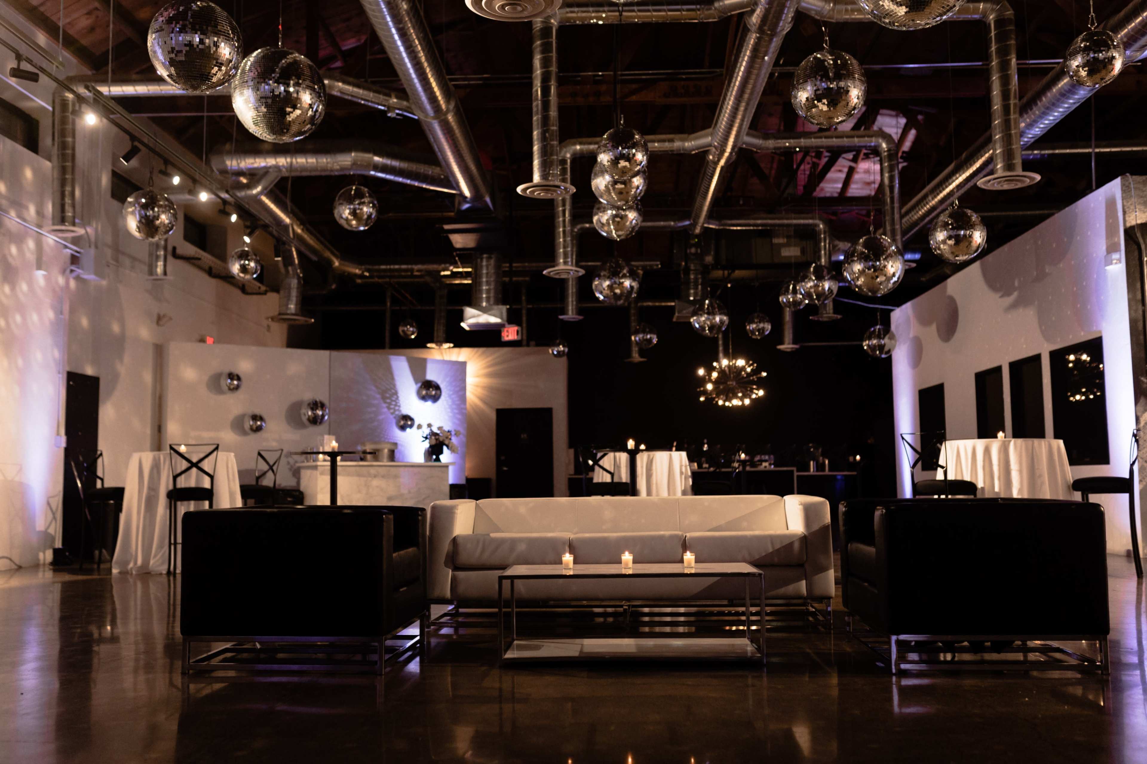 The image shows a sleek event space with modern furniture, metallic decor, and numerous hanging disco balls reflecting light against a dark backdrop.