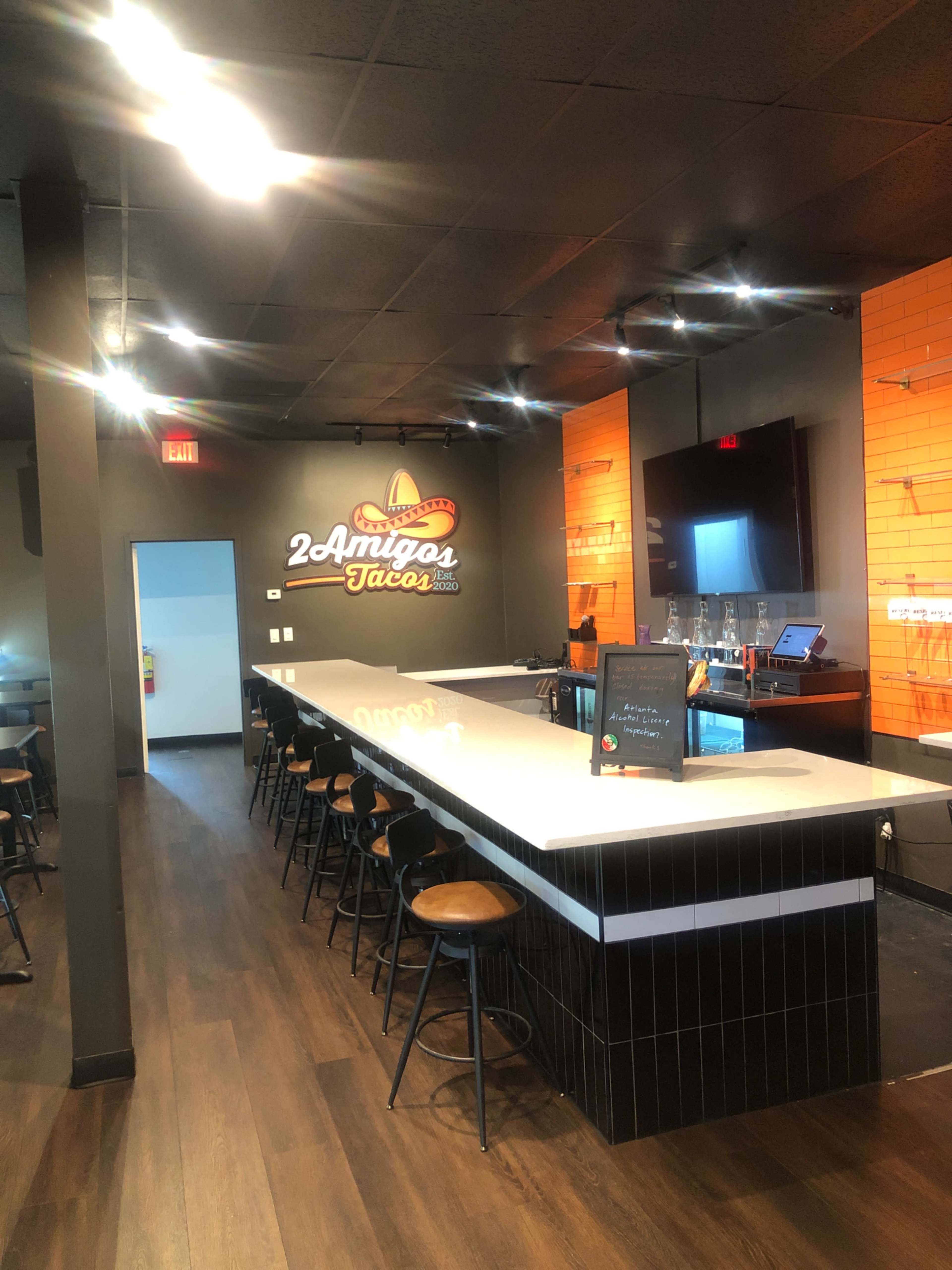 The image shows a modern taco restaurant interior featuring a long white bar counter with wooden stools, orange accent walls, and a chalkboard menu.