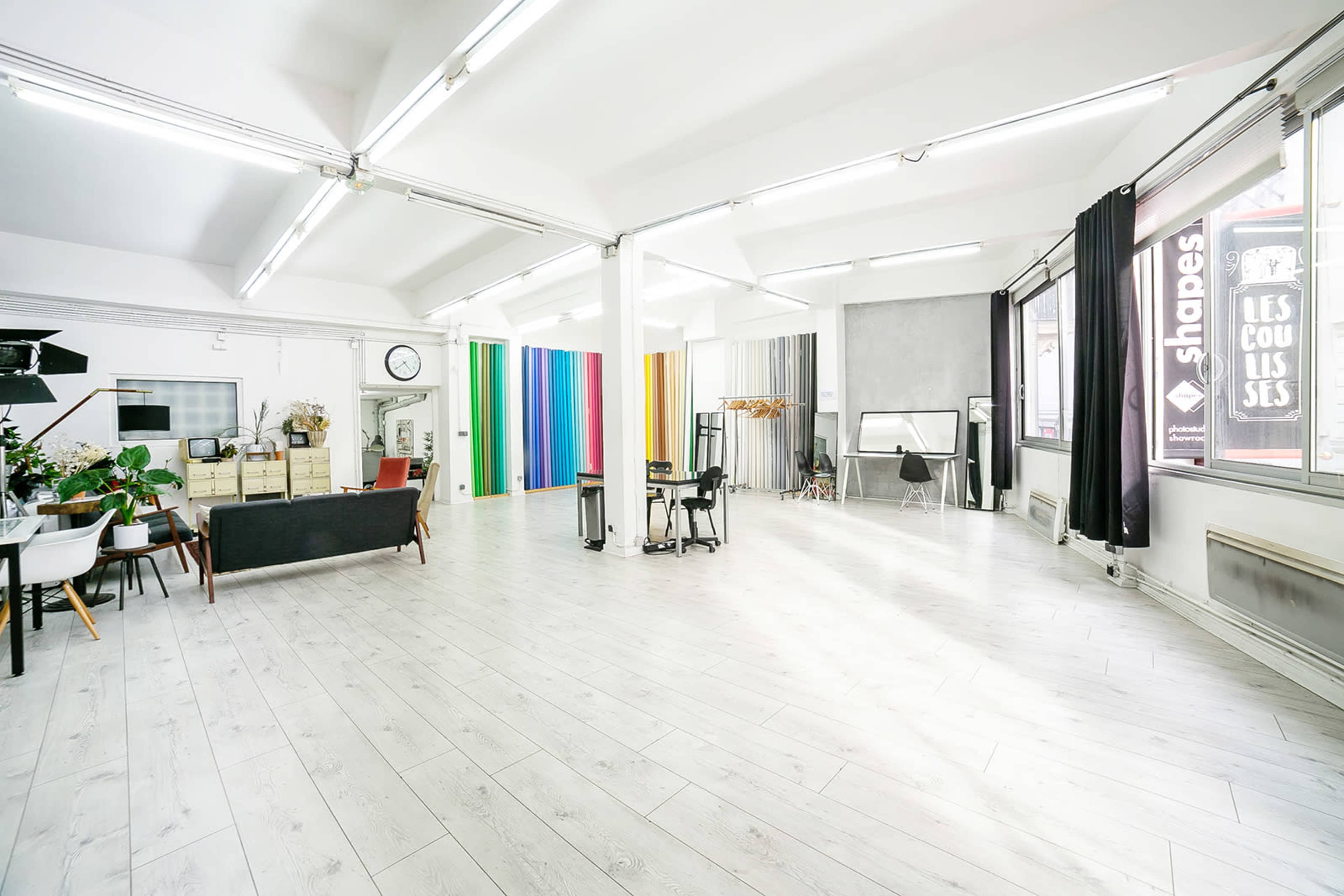 The image shows a spacious studio with white wooden flooring, featuring colorful backdrops, a seating area with a sofa, and various photography equipment.