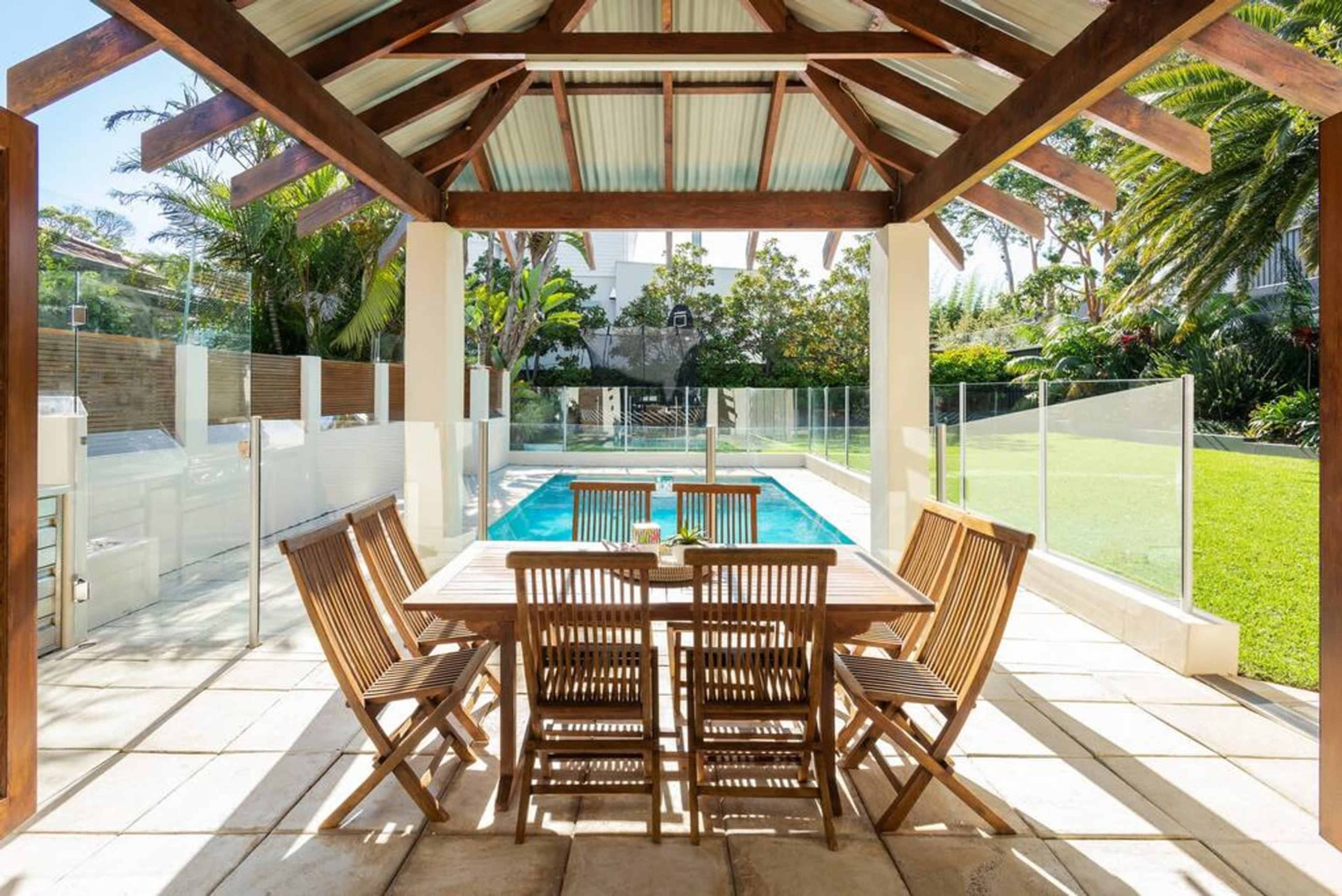 A wooden dining table with chairs is set under a covered patio overlooking a swimming pool and landscaped garden.