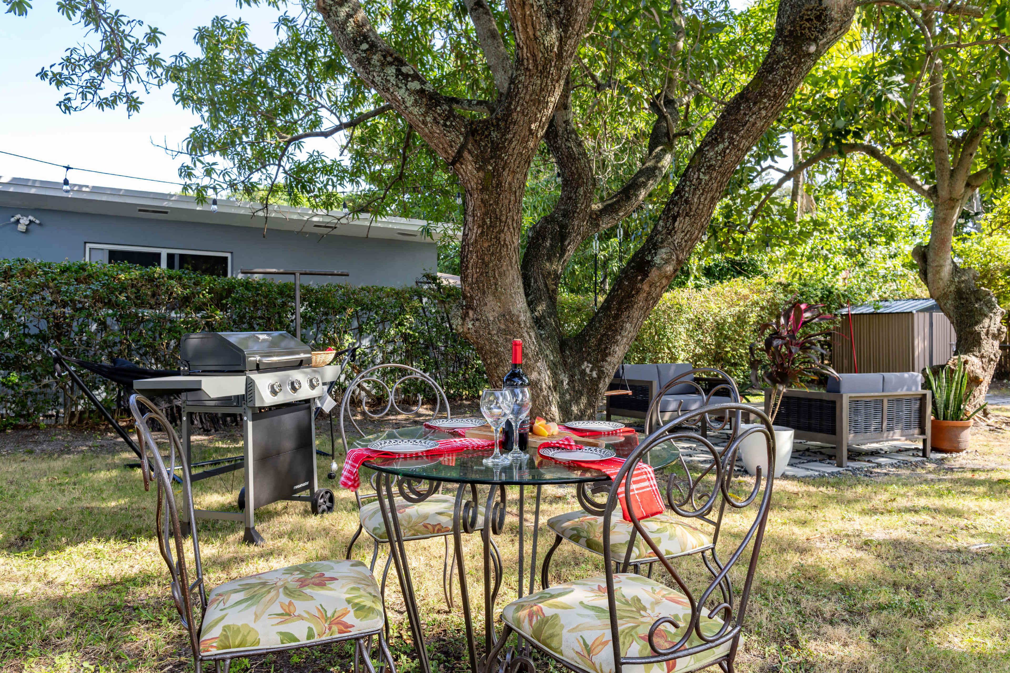 A glass dining table with four chairs is set up in a backyard under a tree, while a grill is positioned nearby and seating areas are visible in the background.