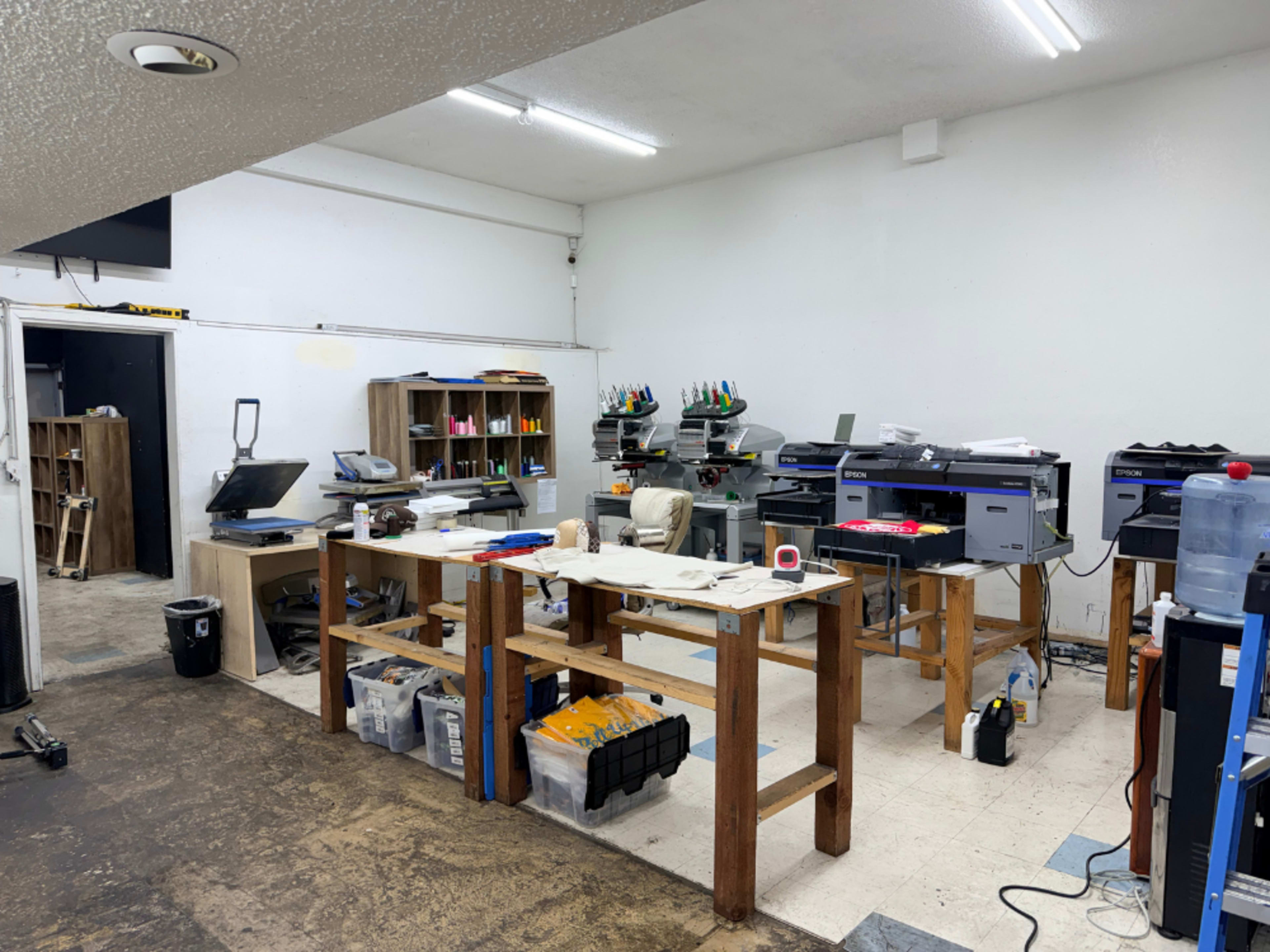 The image shows a printing workspace with multiple printers, wooden tables, and various office supplies organized in containers.