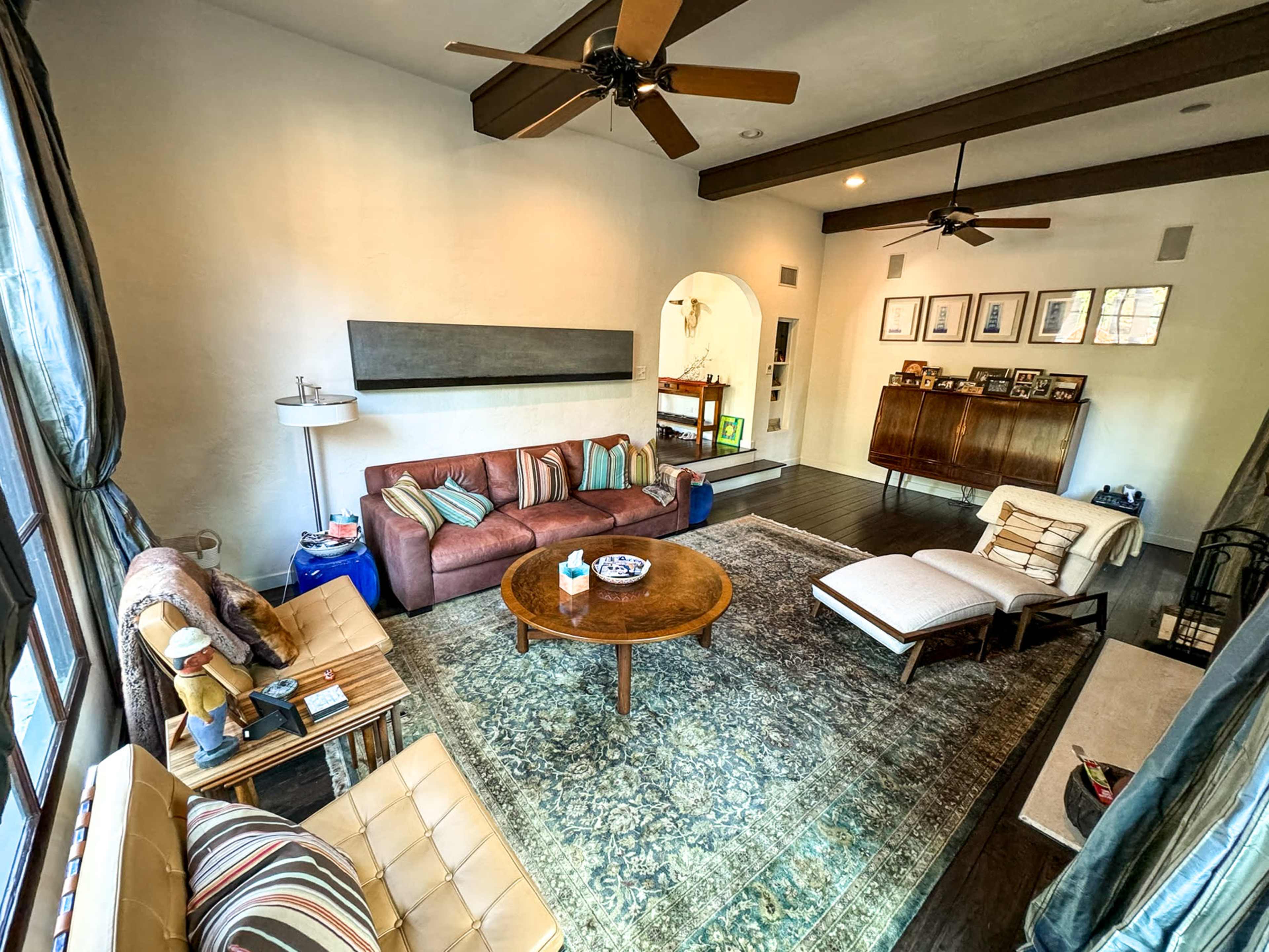 The living room features a central round wooden table surrounded by various seating options, with a large area rug and decorative wall art.