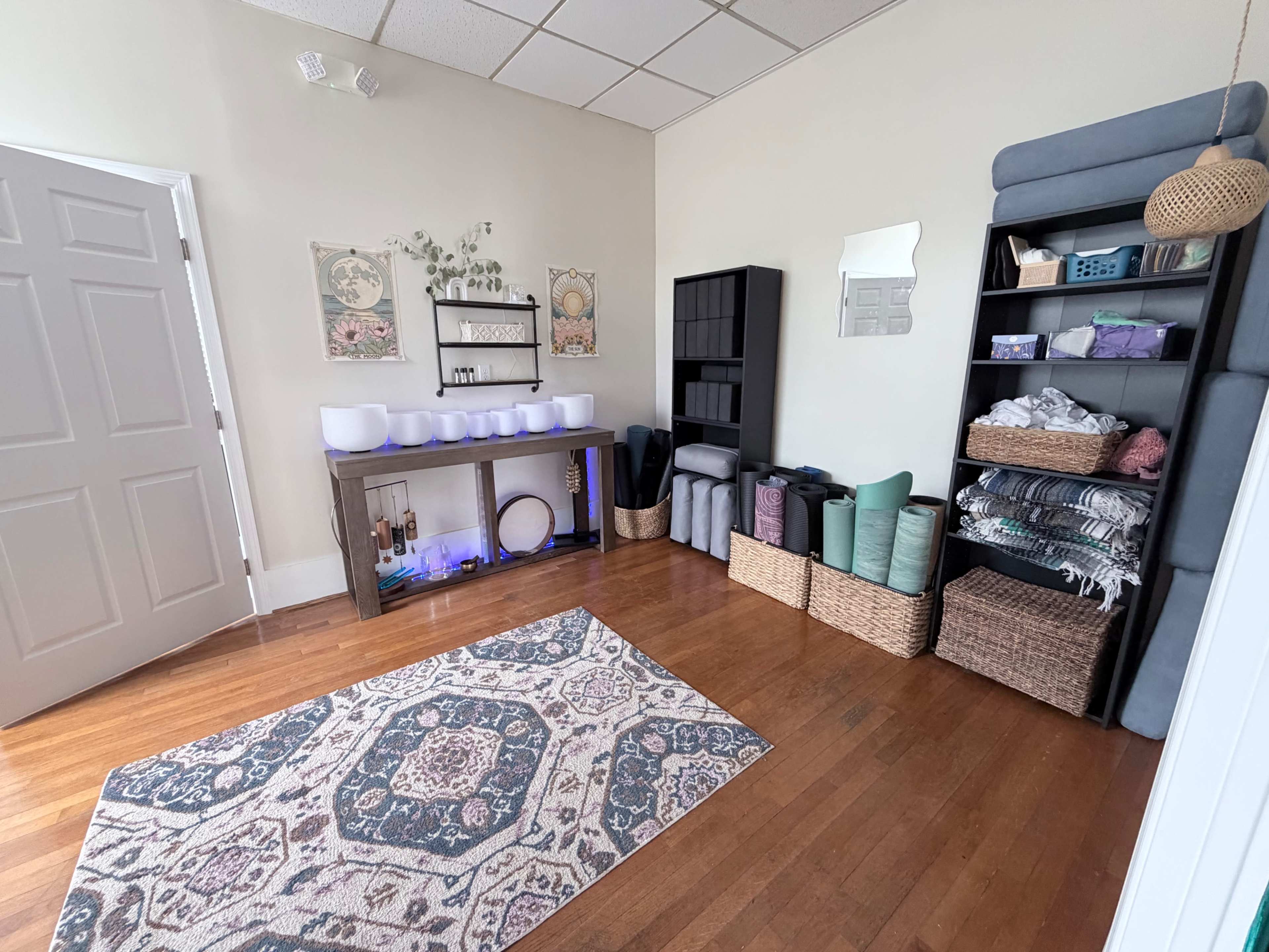 The image shows a calm, organized room with wooden floors, shelves filled with baskets and yoga mats, a console with sound bowls, and wall decorations.