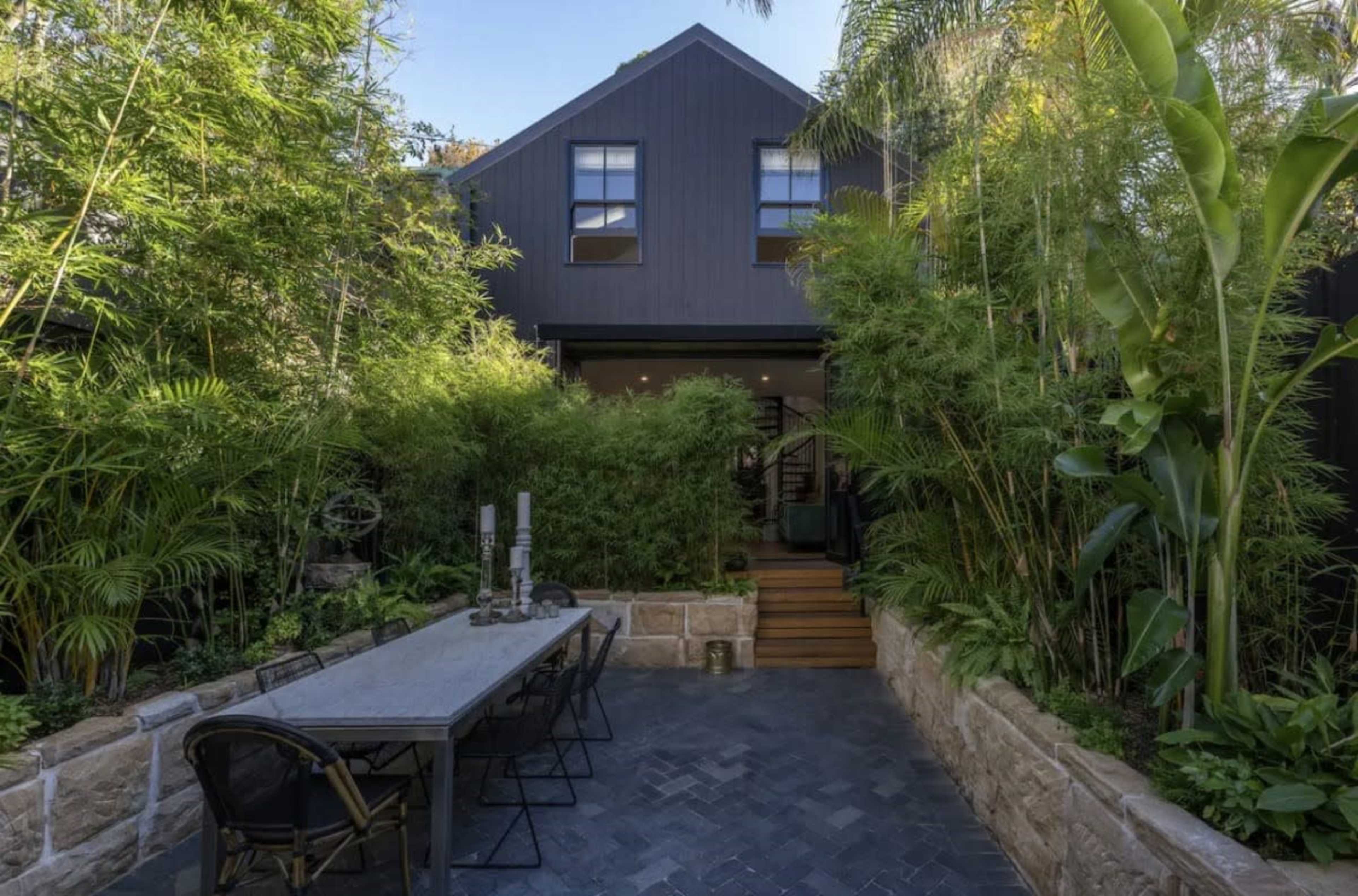A black house surrounded by dense greenery, with a stone patio and a large dining table set in front.
