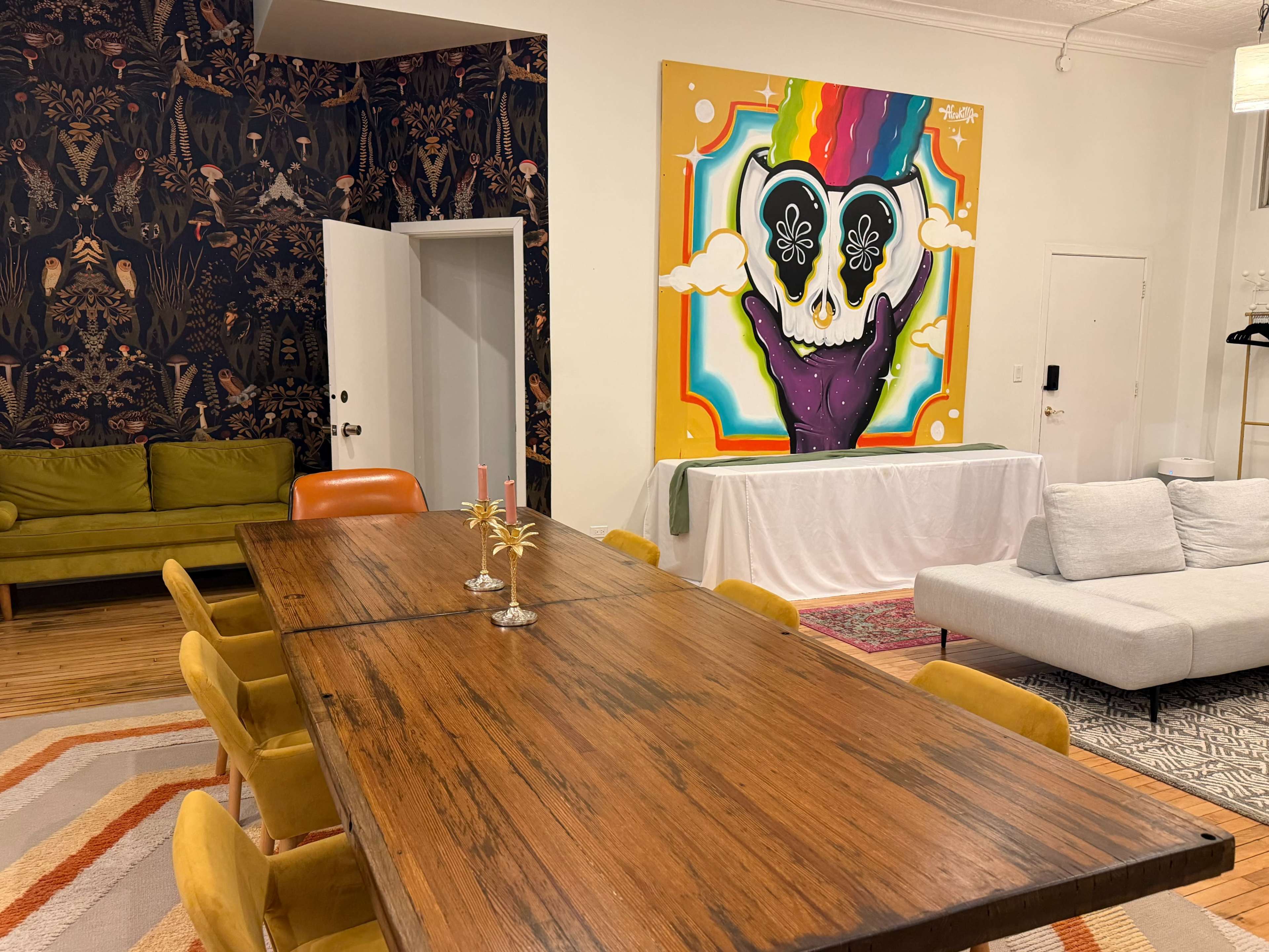 The image shows a brightly decorated room featuring a large wooden dining table surrounded by orange and yellow chairs, with a vibrant wall mural of a skull and colorful design.
