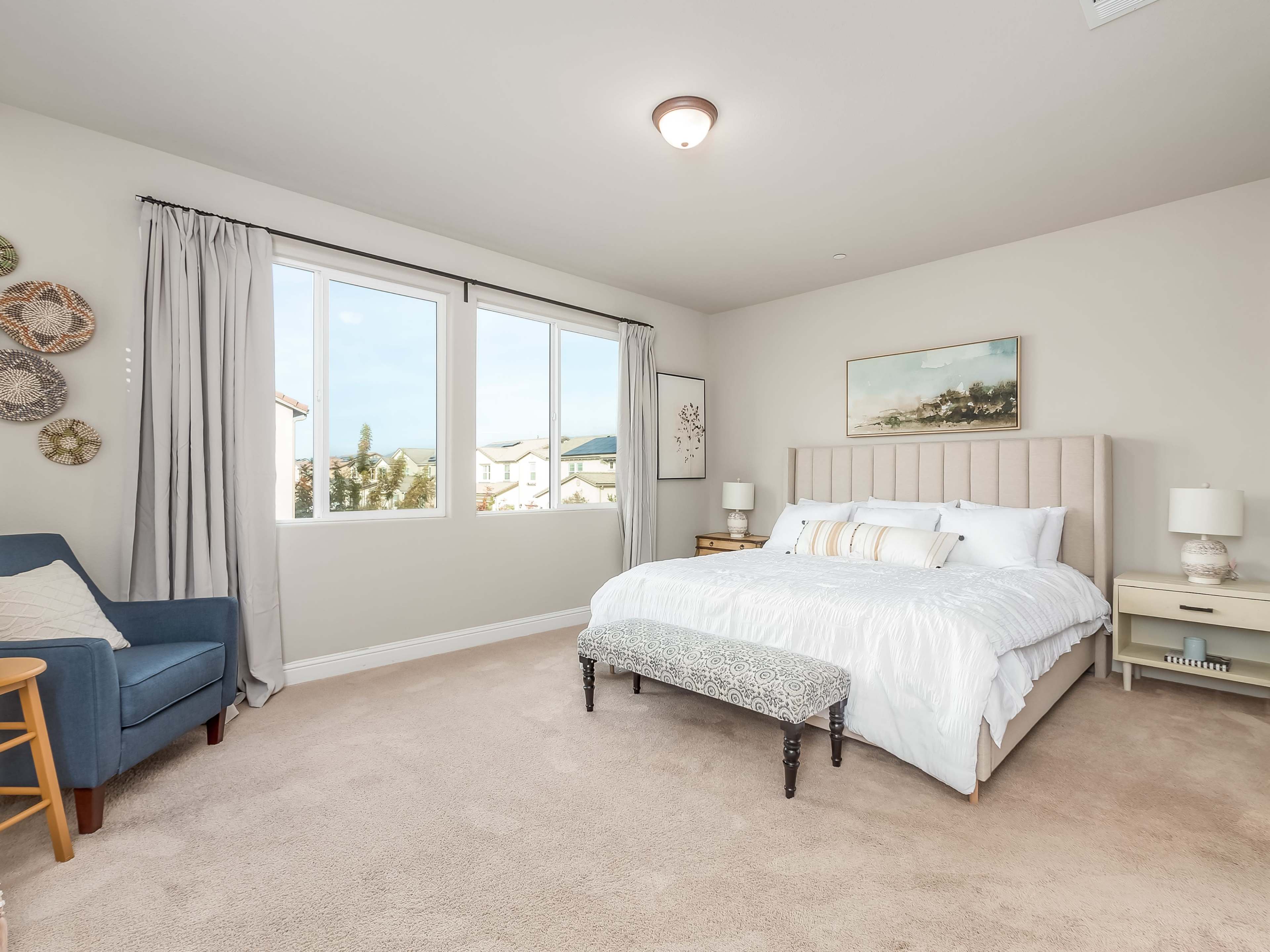 The bedroom features a large bed with white linens, a bench at the foot, a seating area with a blue chair, and windows providing natural light.