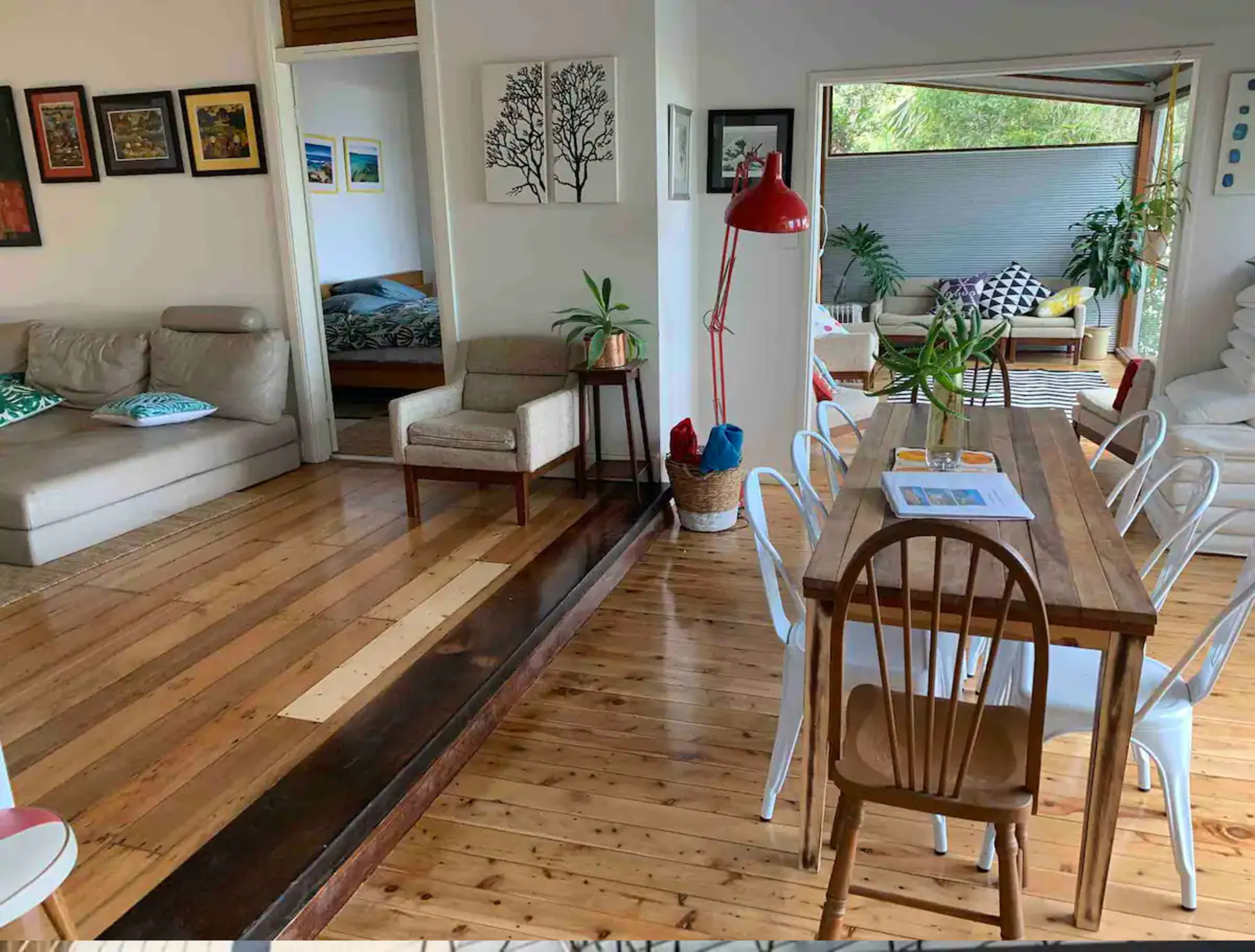 An interior space with wooden floors, a dining table surrounded by chairs, and various seating areas, including a sofa and armchair, alongside wall art and plants.