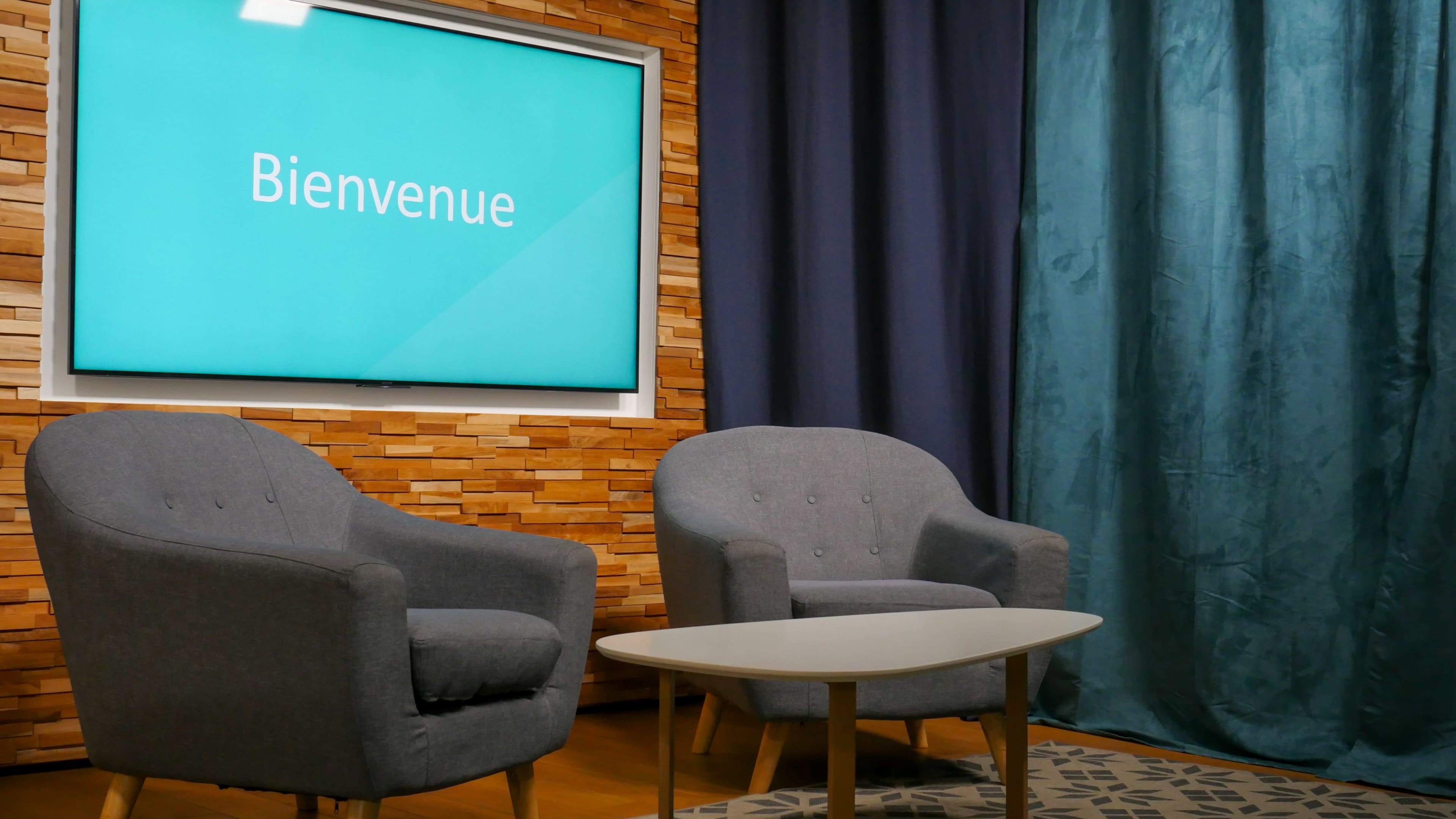 The image shows a modern set with two gray armchairs, a small white table, and a screen displaying the word "Bienvenue" against a wooden wall and dark curtains.
