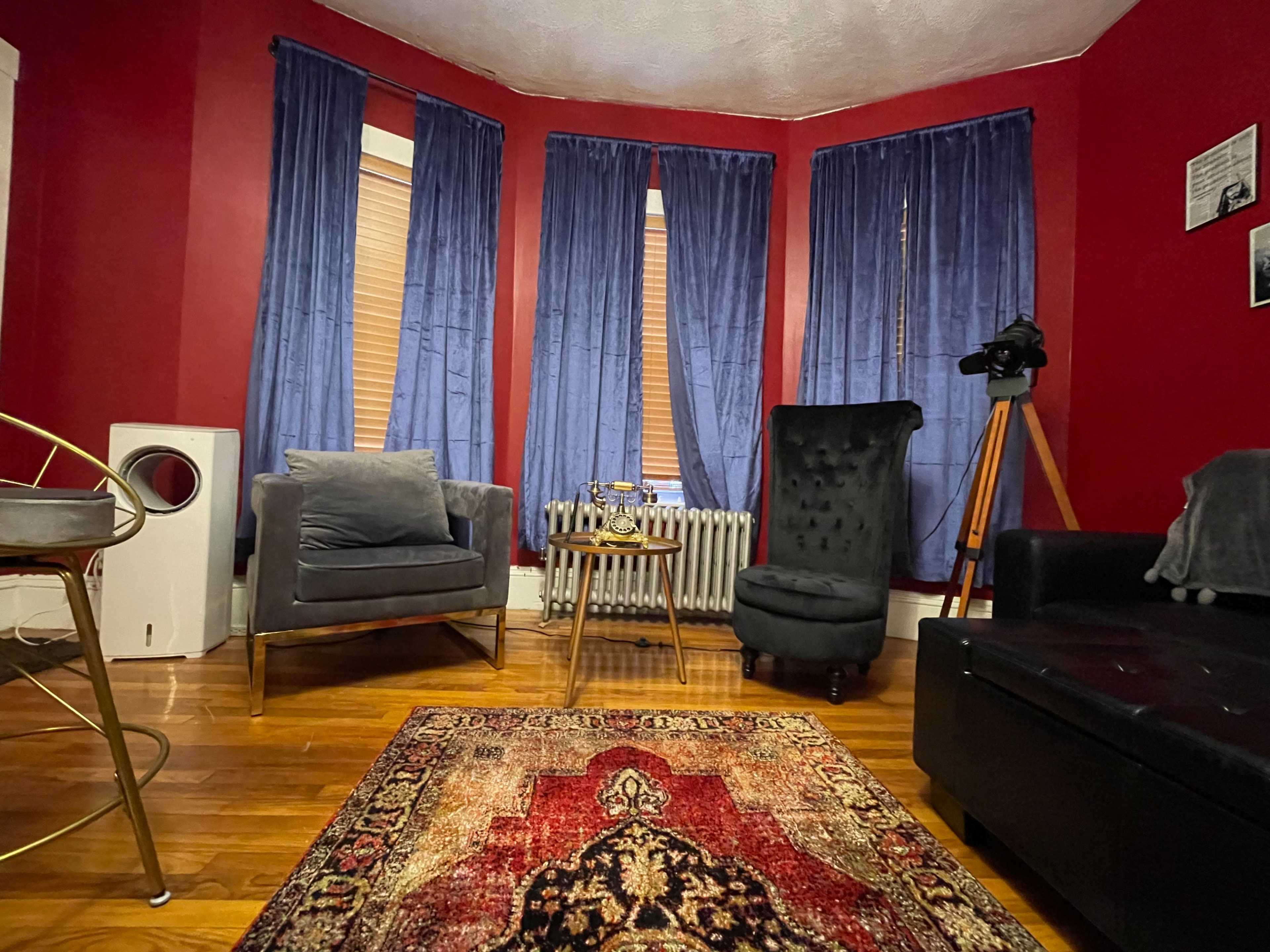 A cozy living room with red walls, two windows covered by blue curtains, and a mix of seating options, including a black sofa, a gray armchair, and a tall black chair beside a small table and an old radiator.