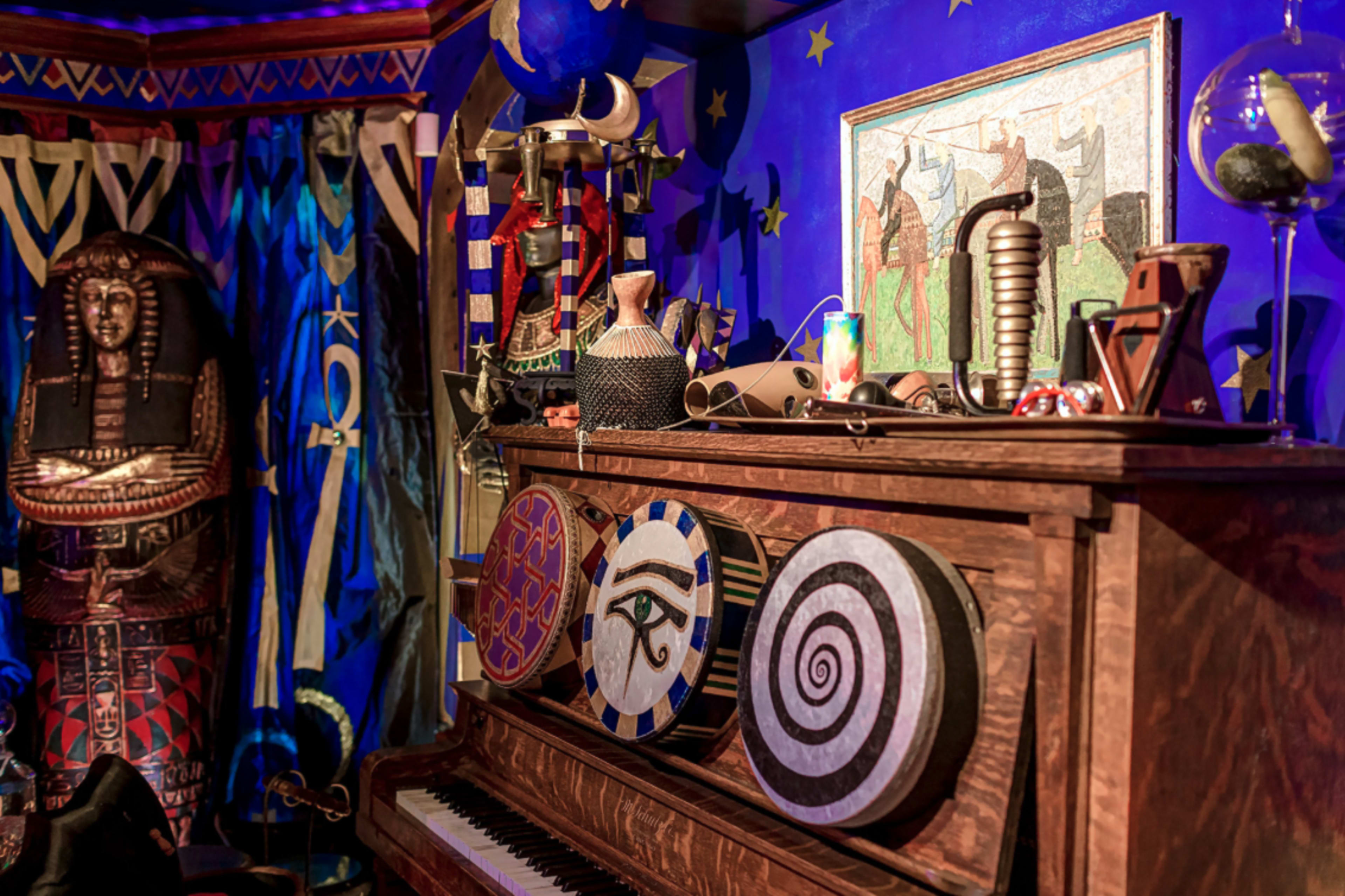 The image features a decorated piano surrounded by various artifacts, including shields and sculptures, in a vividly colored room with star motifs.