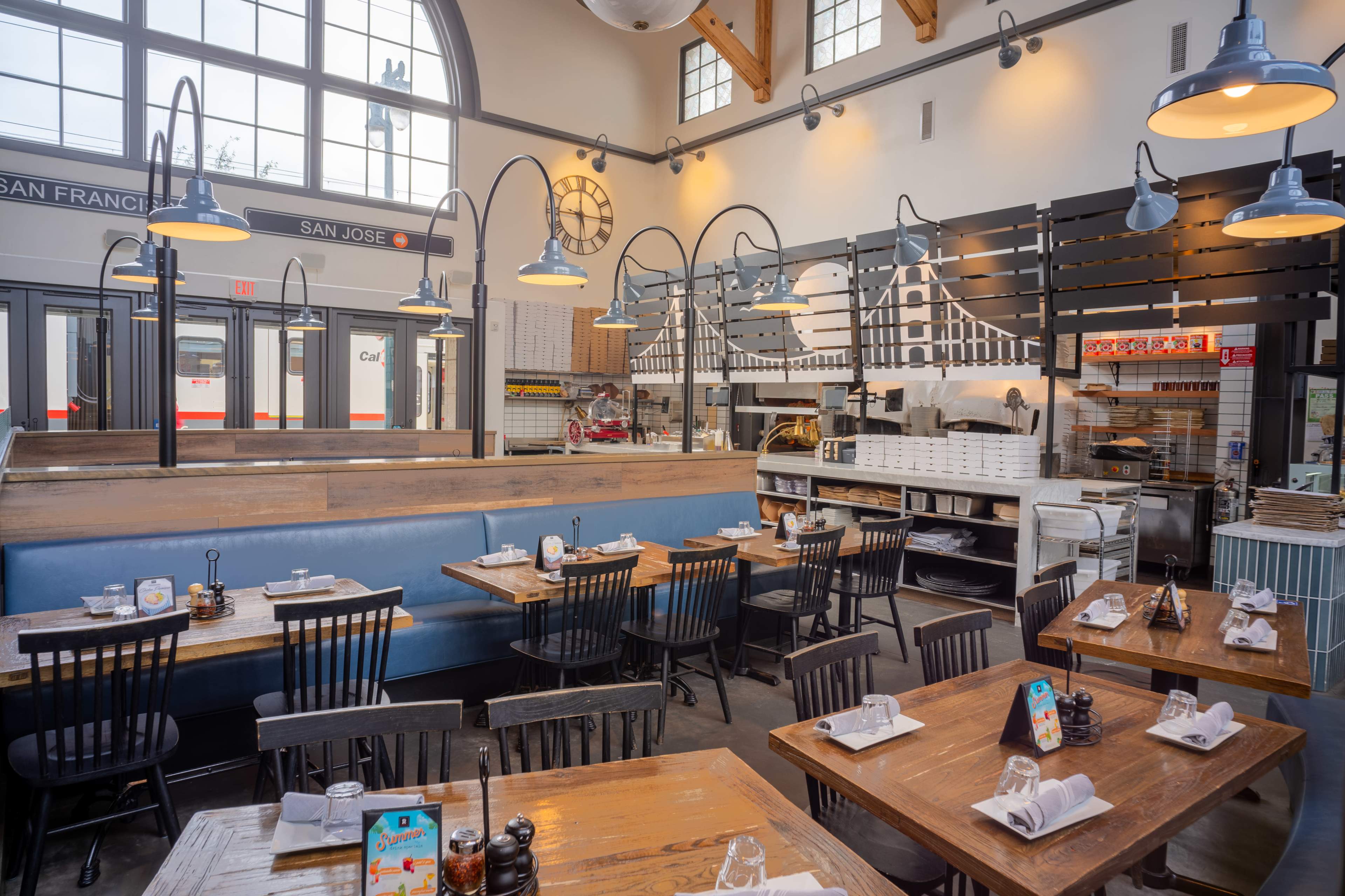 The image shows a spacious restaurant interior with wooden tables and chairs, large windows, and modern lighting fixtures.