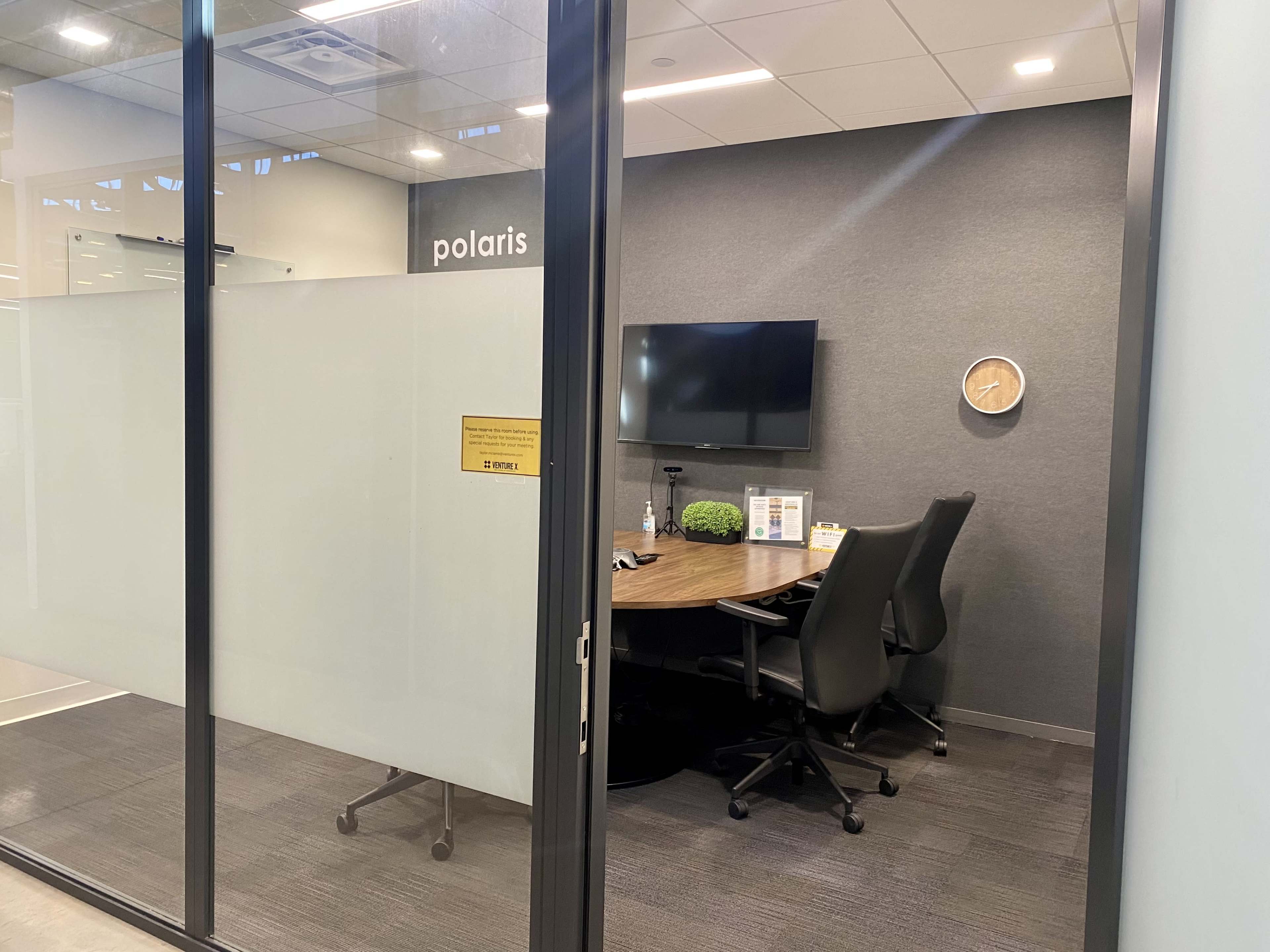 A modern office space with a large desk, two ergonomic chairs, a wall-mounted television, and a clock, enclosed by glass walls.