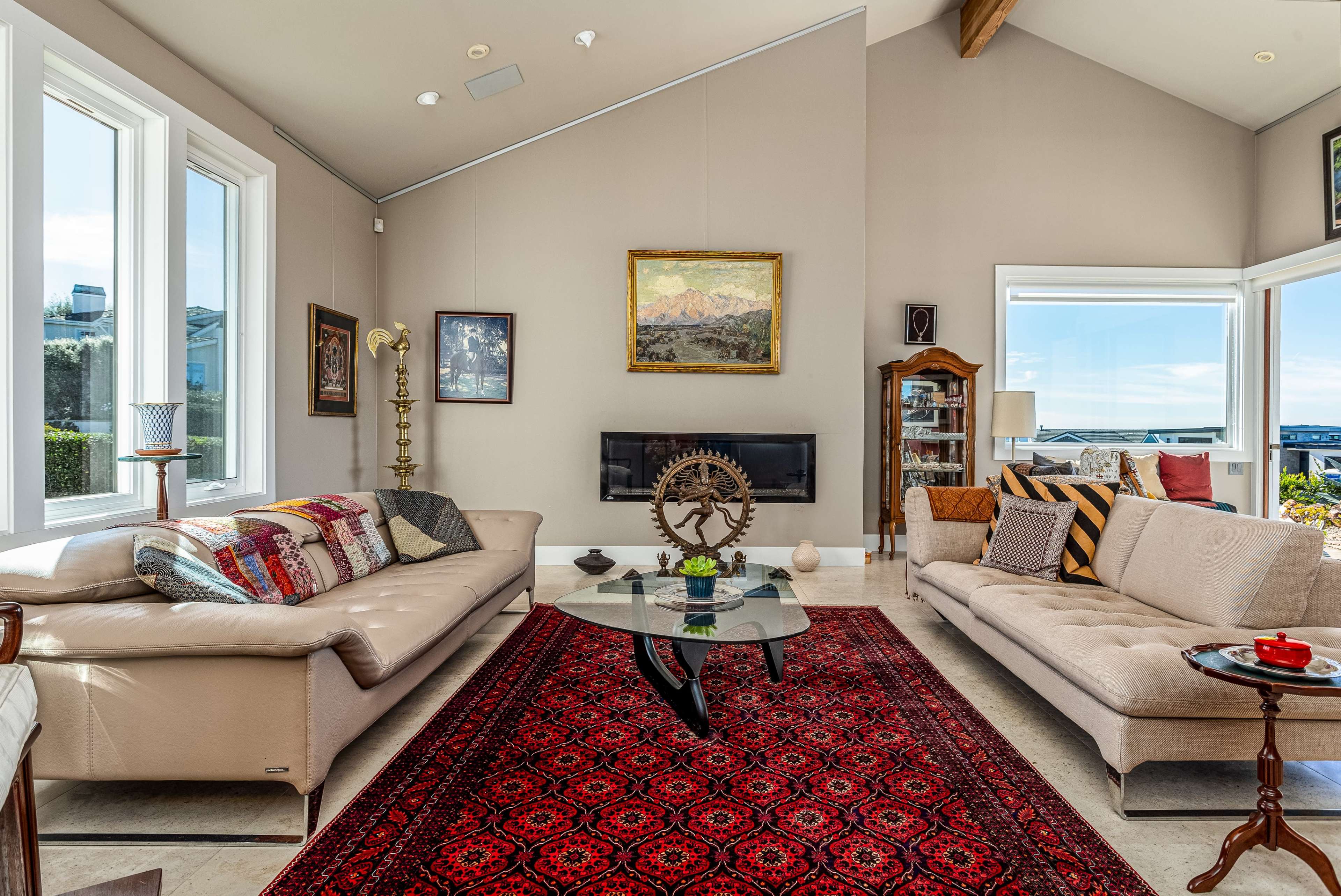 The scene shows a spacious living room with two sofas facing each other, a central glass coffee table, and large windows offering a view of the outdoors.