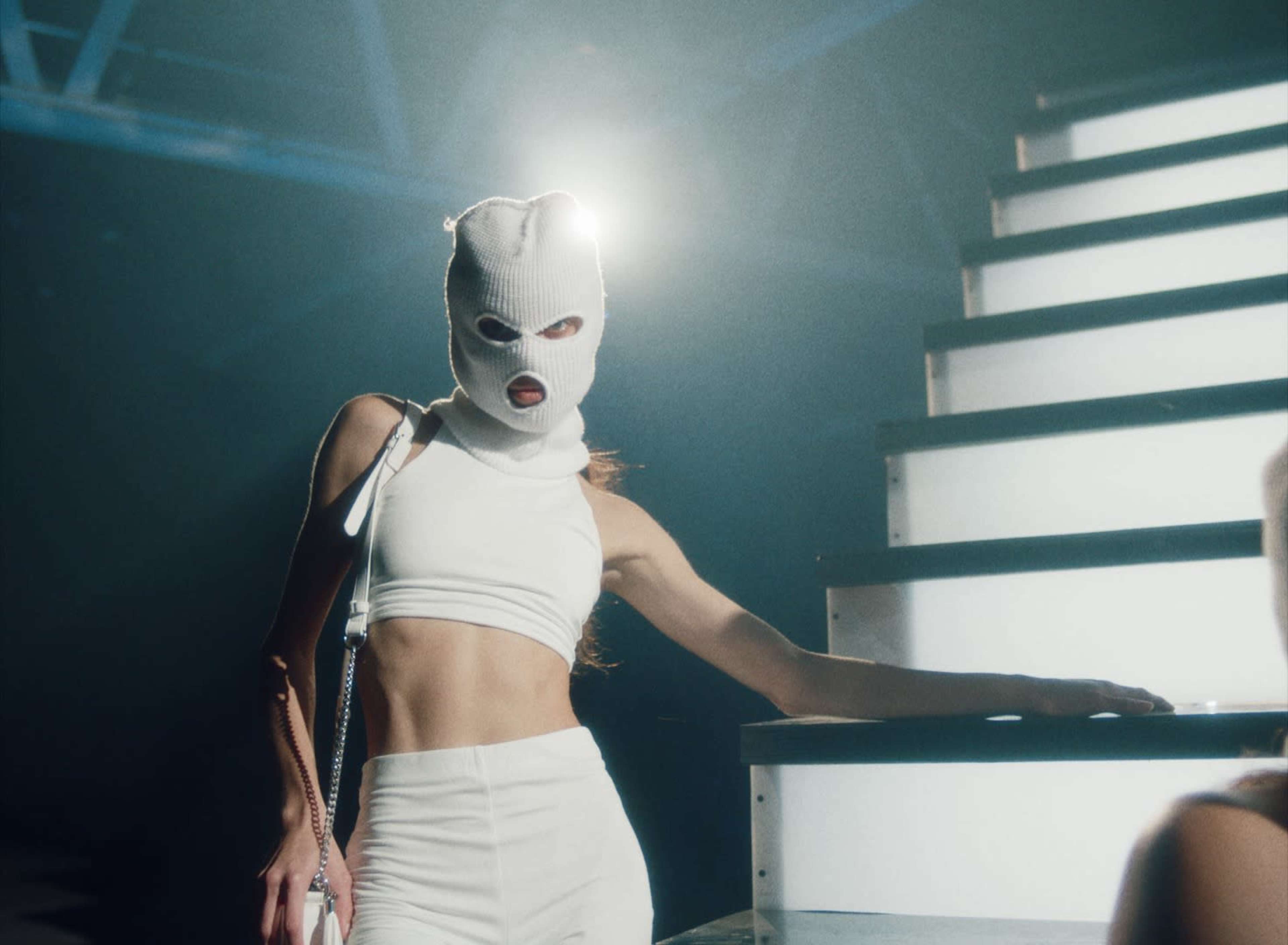 A person in a white outfit and a knit mask poses on a staircase under bright lights.