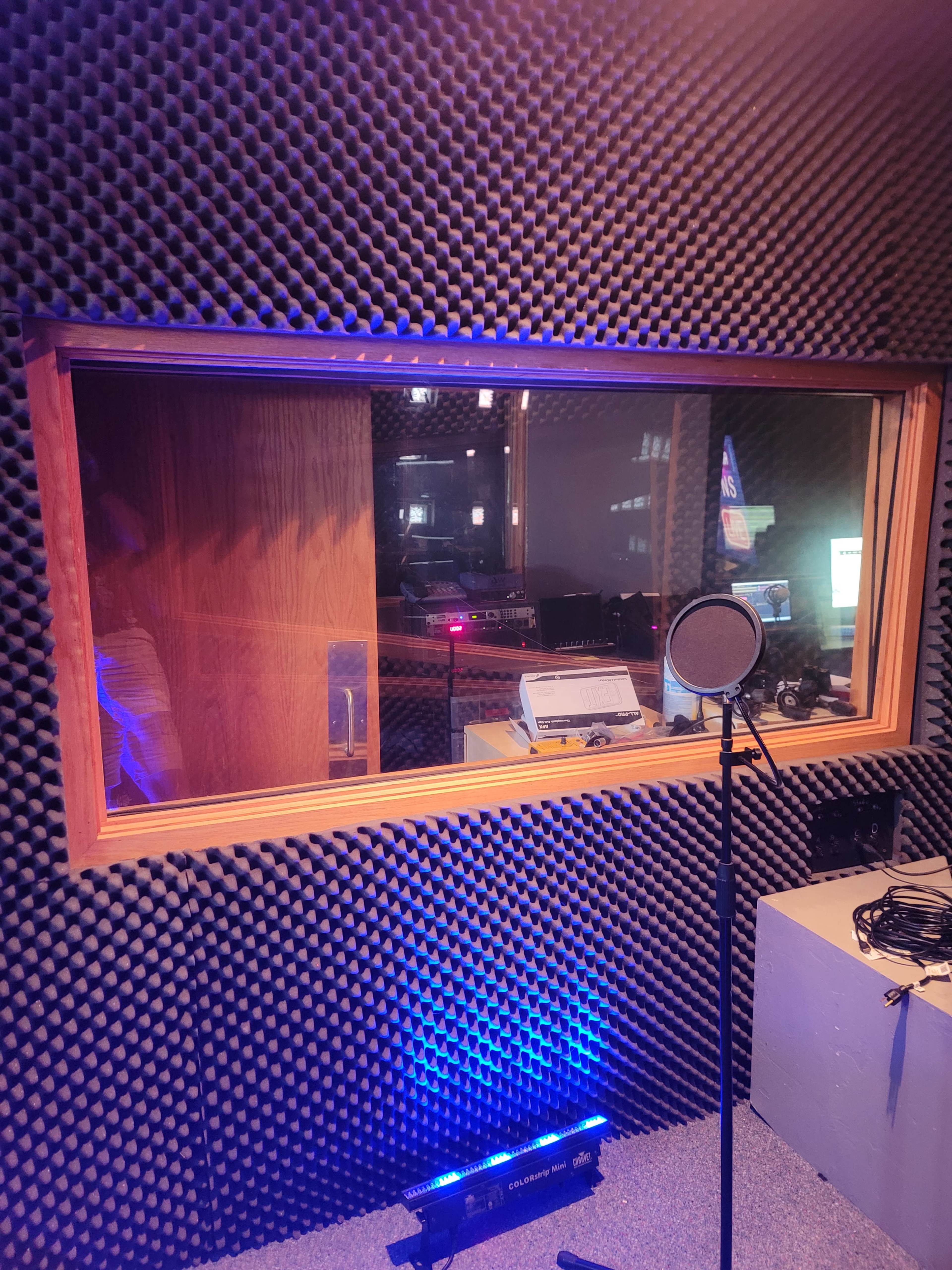 A soundproof recording studio with a microphone in the foreground and equipment visible through a window.