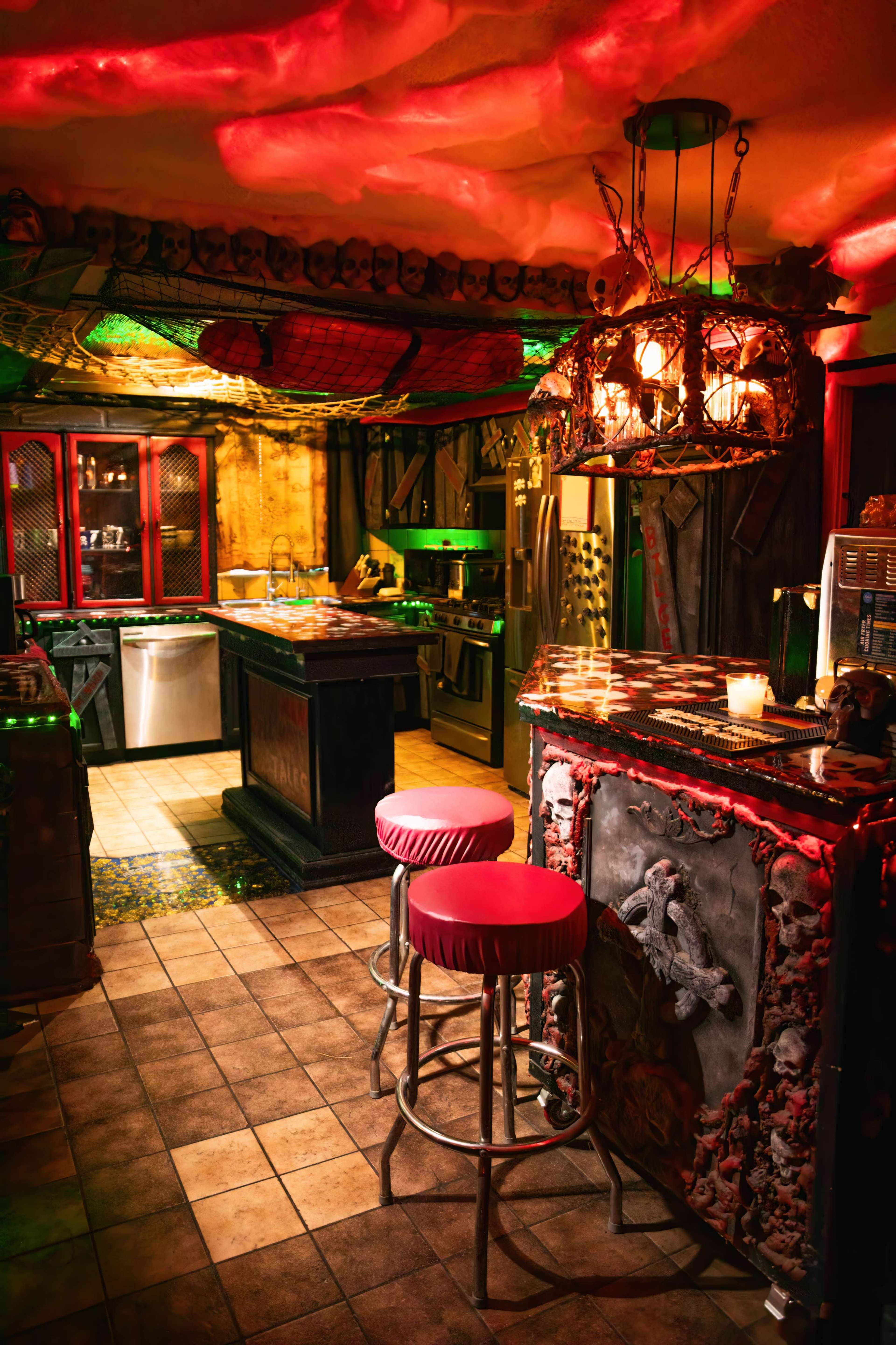 The image shows a dimly lit kitchen with a bar area featuring red stools, ornate decor, and a mix of modern and rustic appliances.
