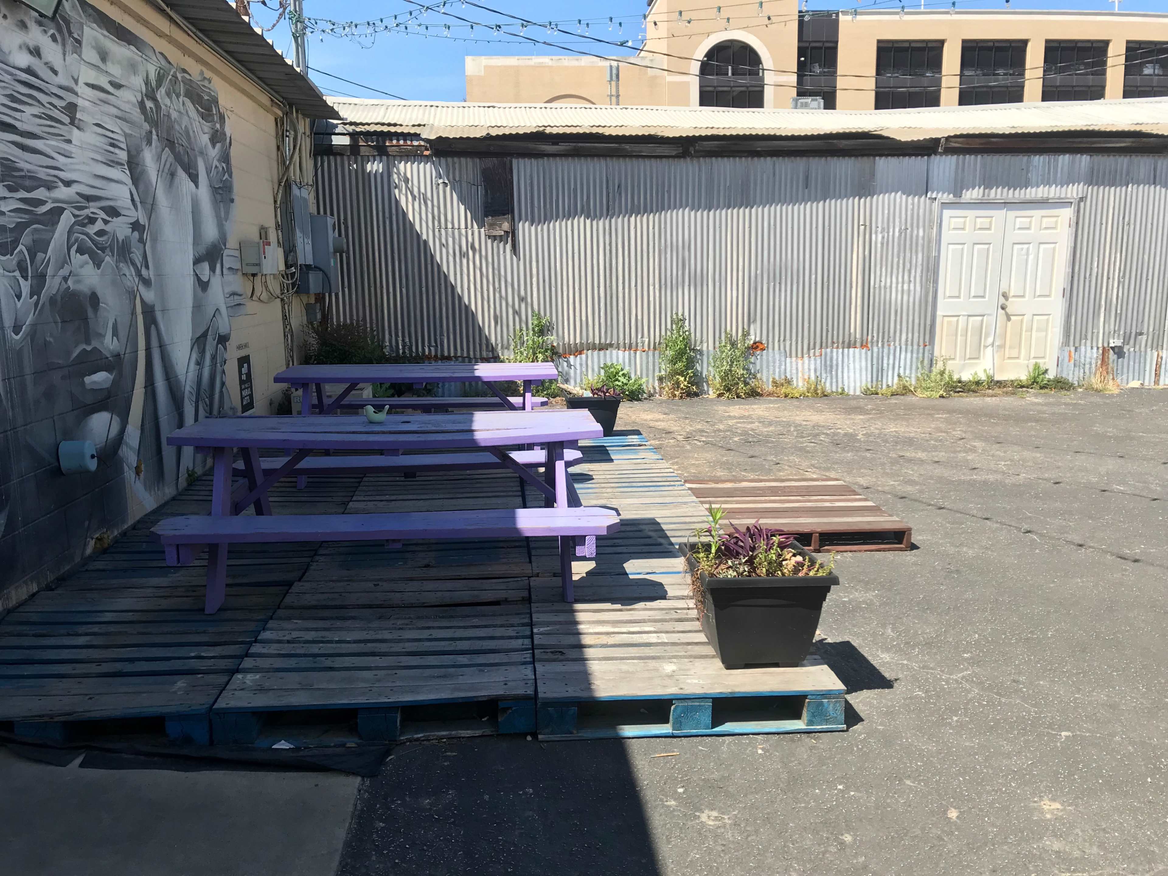 A fenced outdoor area with purple picnic tables on wooden pallets and a corrugated metal backdrop.